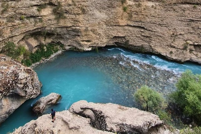 Живописное ущелье с бирюзовой рекой Аксу в районе Шымкента, Казахстан, протекающей между скалистыми берегами. Вода прозрачная, позволяющая увидеть дно с галькой. На переднем плане человек стоит на краю скалы, наслаждаясь видом. Скалы окружают реку с обеих сторон, а небольшая растительность добавляет зелёные акценты. Картина передаёт атмосферу природной красоты и спокойствия.