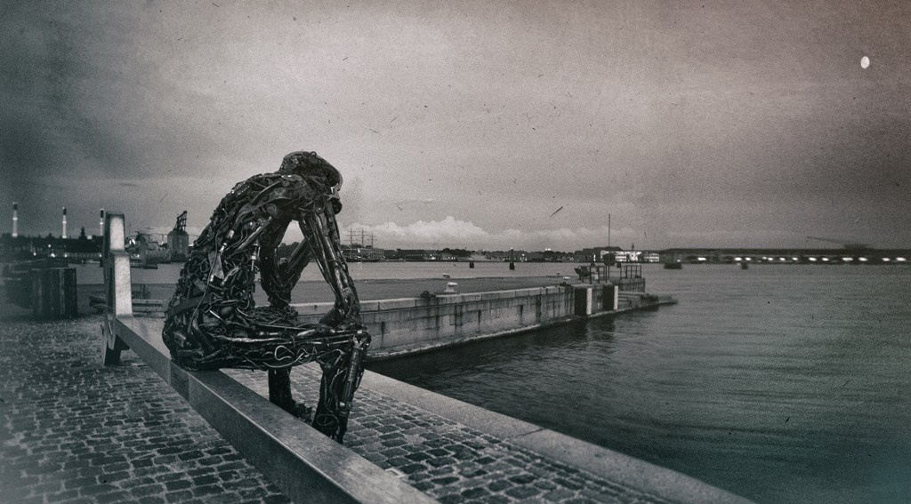 Черно-белая фотография: скульптура из металлических цепей в виде сидящего человека на краю причала, гавань и вода на фоне, зернистая текстура, луна на небе, атмосферное фото