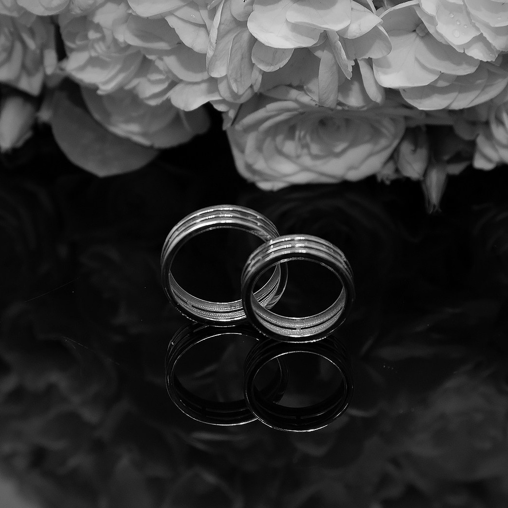 черно белое фото обручальных колец на темном зеркальном фоне рядом с букетом цветов 