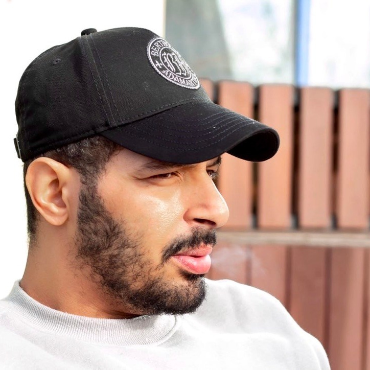 "man in a black baseball cap and white shirt, looking thoughtfully outdoors beside a wooden fence"