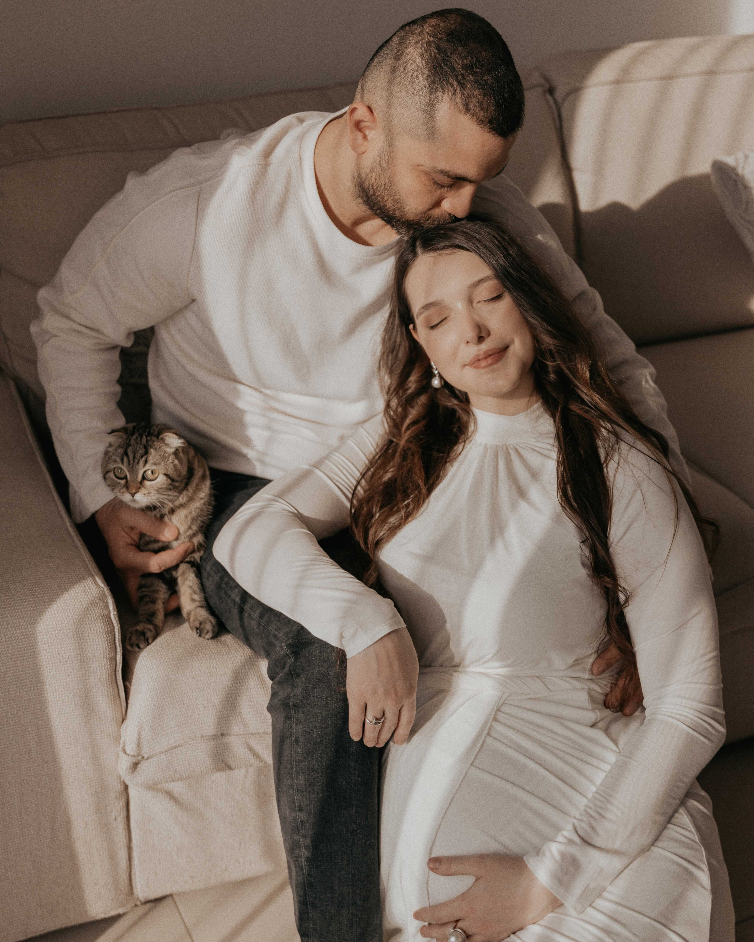 A couple embraces on a beige couch in a warmly lit room. The man kisses the woman's forehead as she rests her hand on her pregnant belly. A grey tabby cat sits calmly beside them. The scene exudes love and anticipation