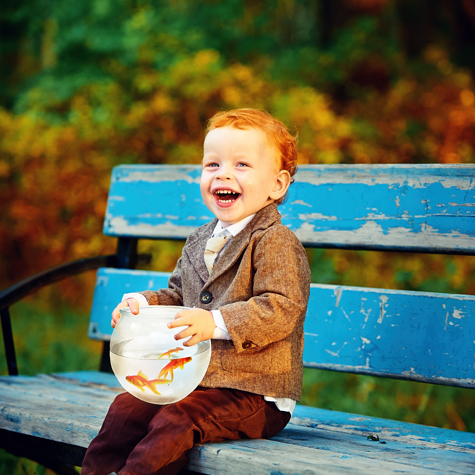 Рыжий мальчуган сидит в парке на синей лавочке с аквариумом в руках и заразительно смеётся