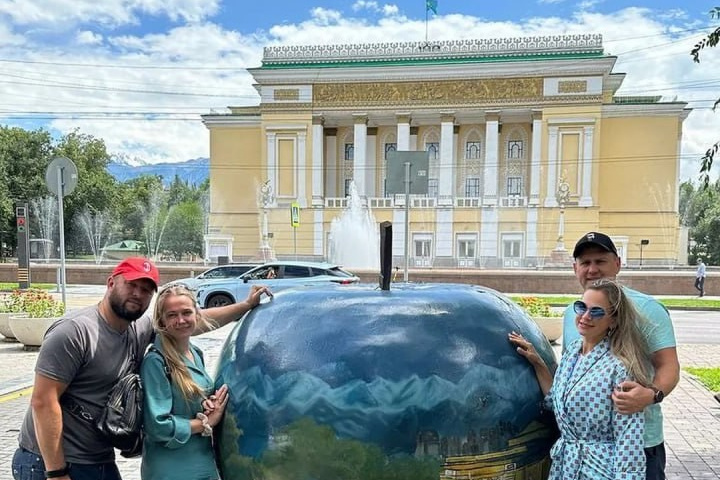 Человек позирует на фоне бирюзового озера Иссык в районе Алматы и гор. Он стоит на большом камне, одет в ярко-зеленую толстовку, черные штаны и светлую обувь, а также оранжевую панаму. В руках держит очки. За ним виднеются деревья, лесистые склоны и горные вершины под облачным небом. Атмосфера спокойная и природная.