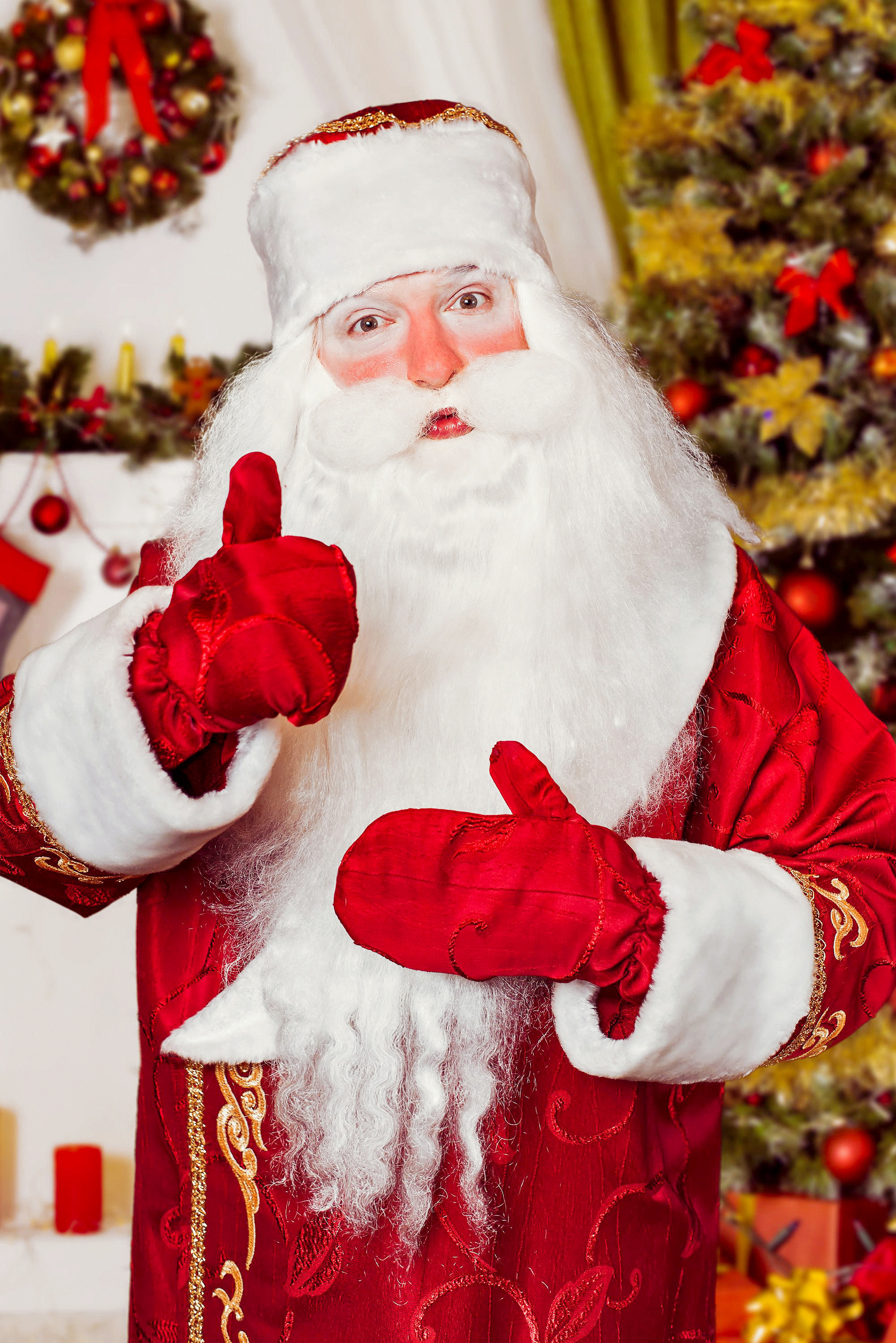 Дед Мороз на фоне новогодней ёлки показывает класс пальцем вверх.