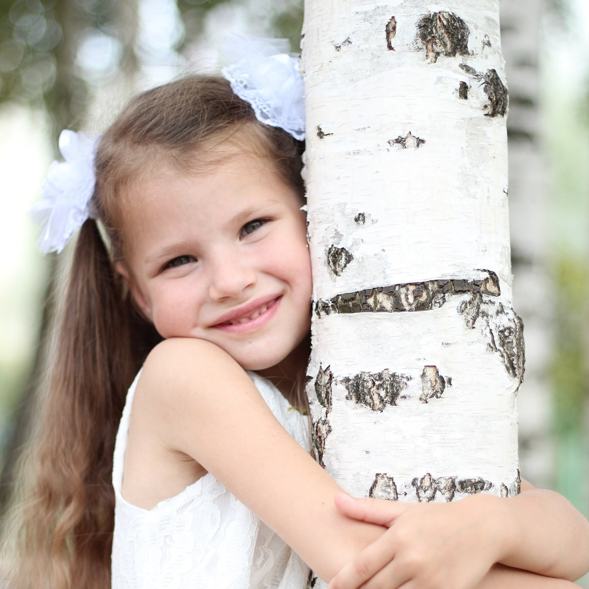 девочка в белом платье с белыми бантами обнимает березу
