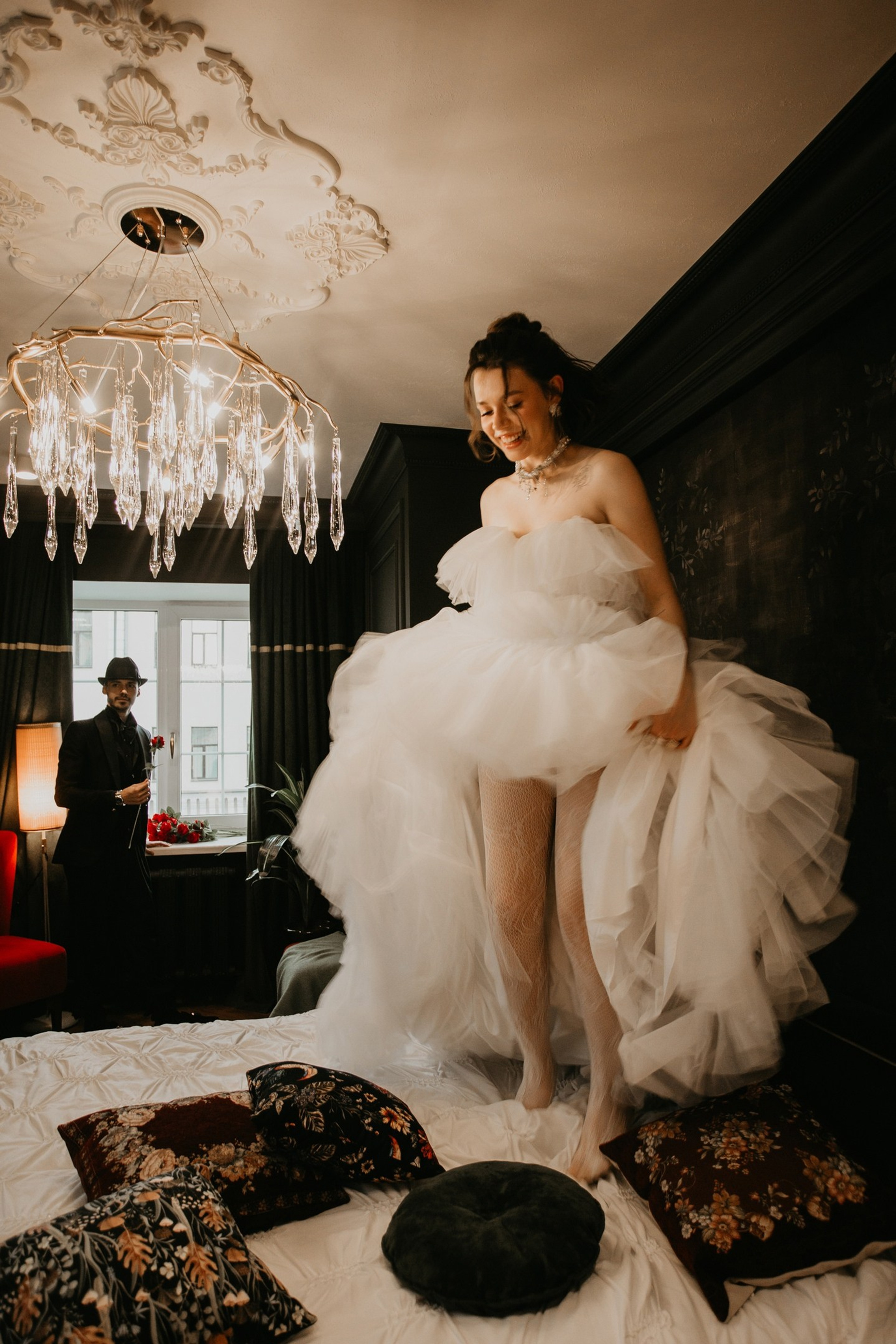 Свадебная фотосессия - невеста прыгает на кровати, а жених на фоне у окна