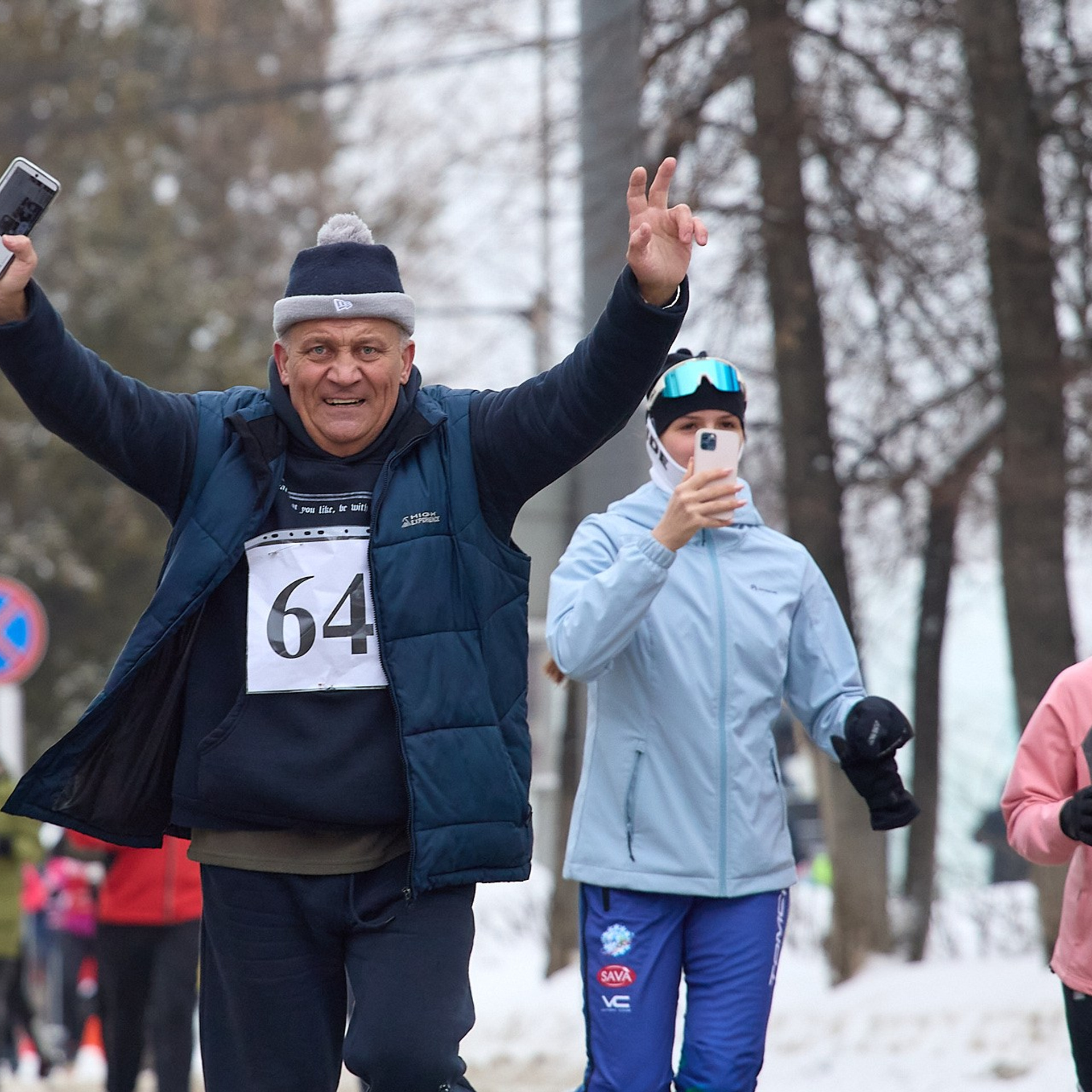 Солидный возрастной мужчина в расстегнутой куртке с номером 64 на груди бежит навстречу впереди всех, подняв руки.