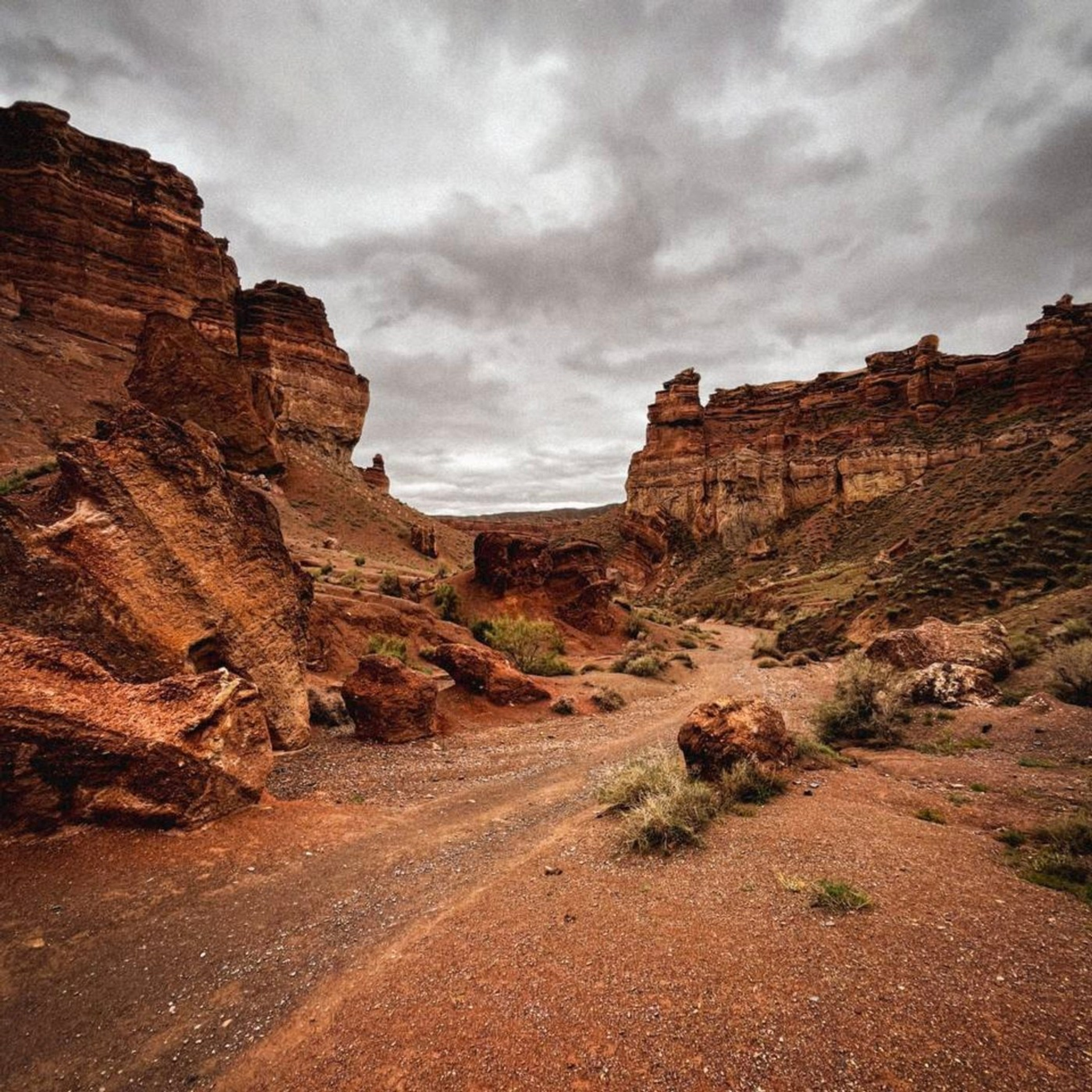 Пейзаж Долины Замков с красновато-коричневыми породами под пасмурным серым небом. На переднем плане видны крупные камни и грунтовая тропа, ведущая вдоль Чарынского каньона. Растительность редкая, подчёркивая суровость и красоту этого природного места.