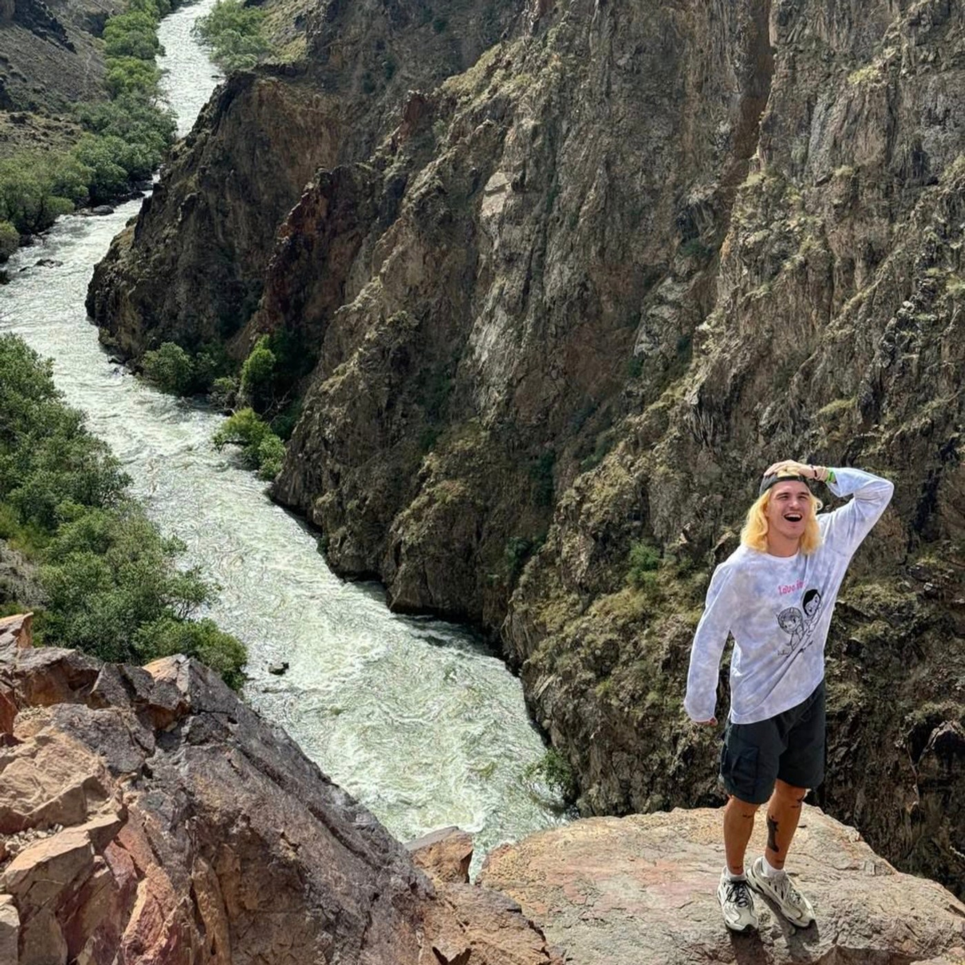 Молодой человек стоит на вершине утеса с видом на глубокий Черный каньон, через которое протекает бурная река Чарын. Скалистые стены ущелья покрыты редкой растительностью, а внизу видны деревья вдоль русла реки. Мужчина выглядит восторженно, позирует с одной рукой на голове.