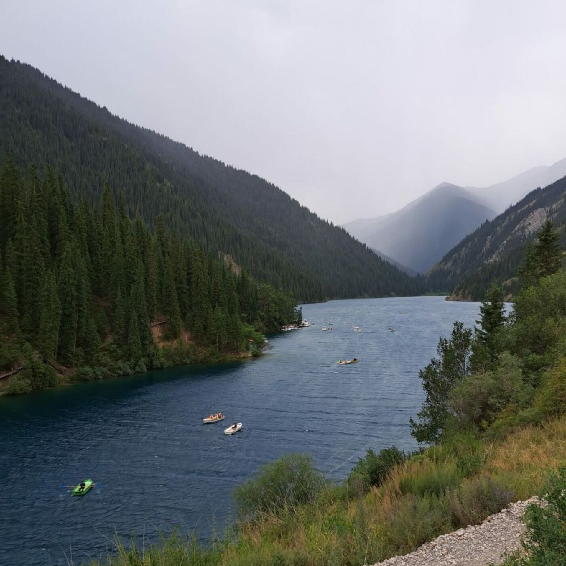 Горное озеро Кольсай, окруженное густым хвойным лесом, под серым облачным небом. Вдоль озера можно увидеть несколько лодок и катамаранов на воде, создающих атмосферу спокойствия и отдыха. Холмы и горы вдали покрыты лесом, а передний план украшен травянистым склоном и гравийной дорожкой.