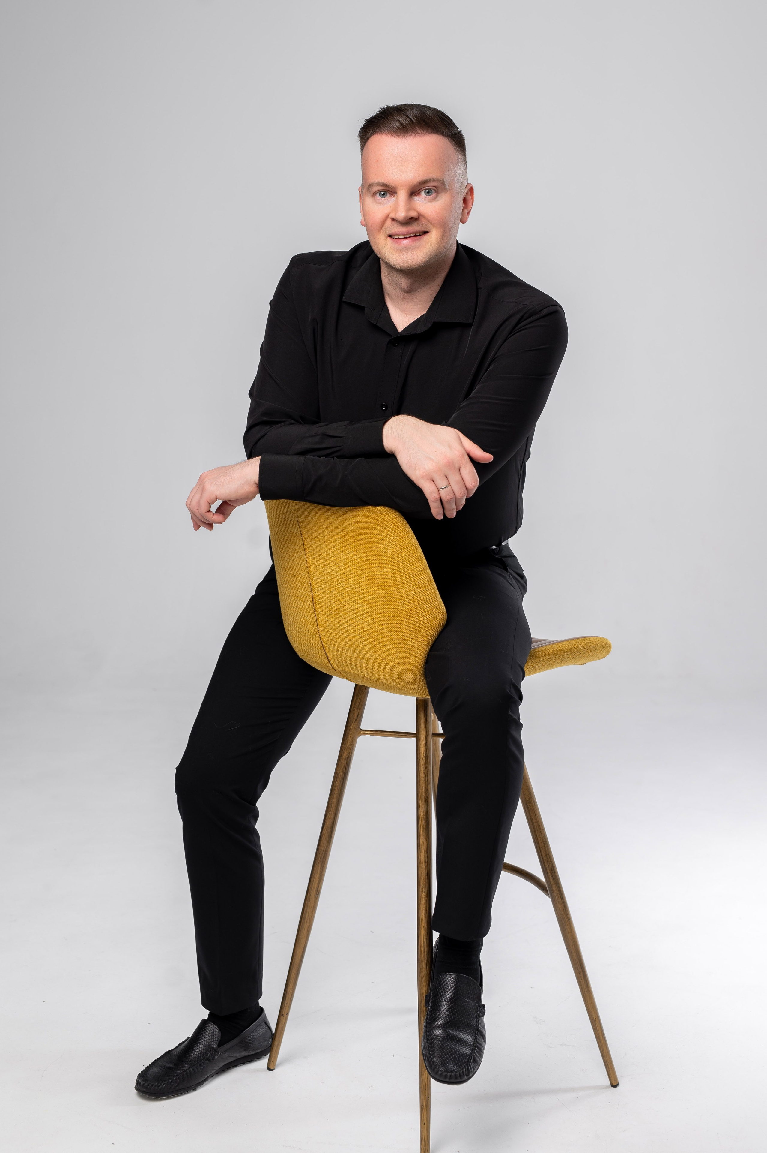 Стильно одетый во всё черное мужчина позирует, сидя на высоком на белом фоне циклорамы в фотостудии СПб во время индивидуальной портретной мужской фотосессии.