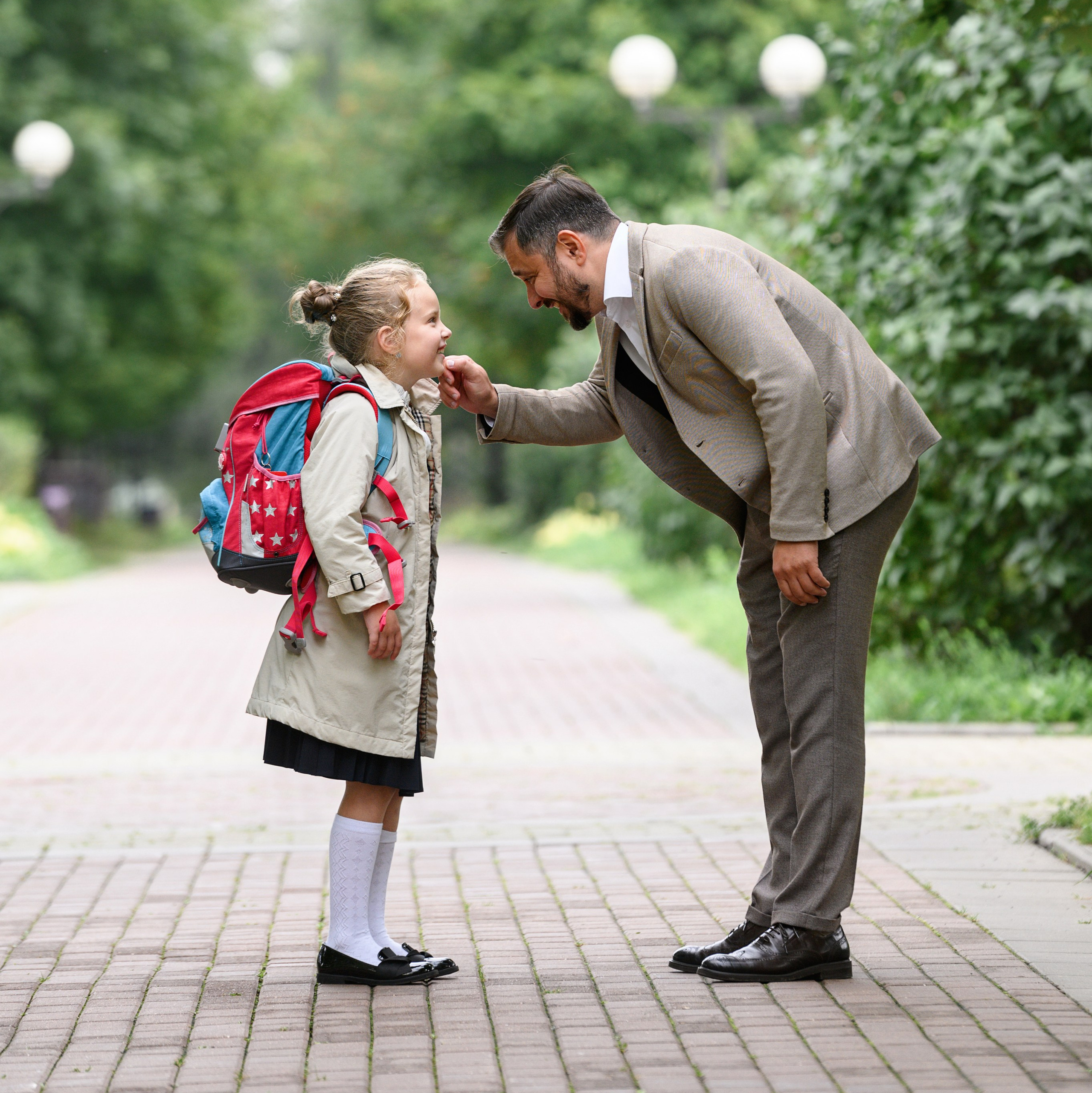 Взрослый наклонился к ребёнку на аллее в парке перед началом учебного дня.