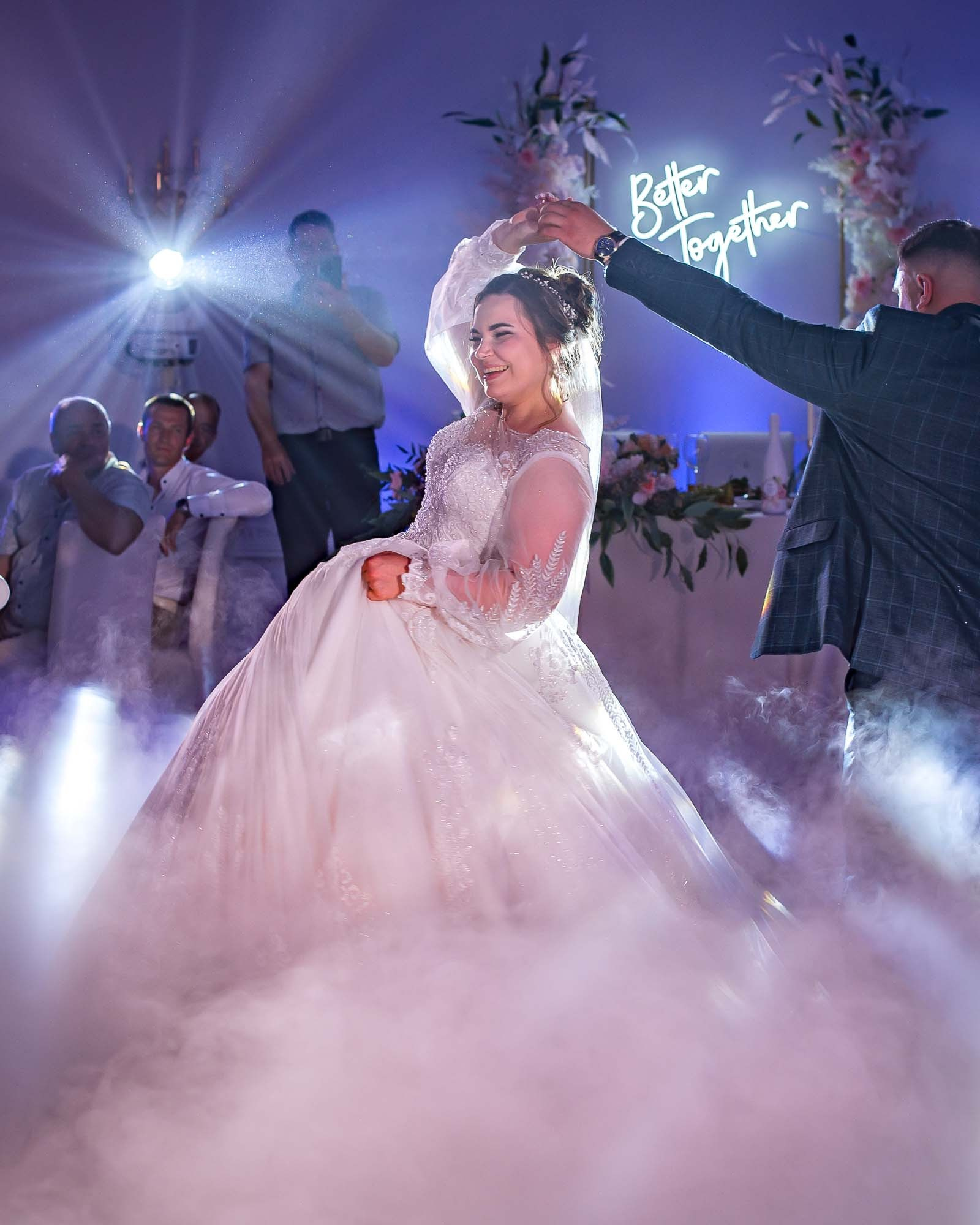 жених танцует с невестой на фоне света софитов, на танцполе тяжелый дым, вокруг за столиками сидят гости