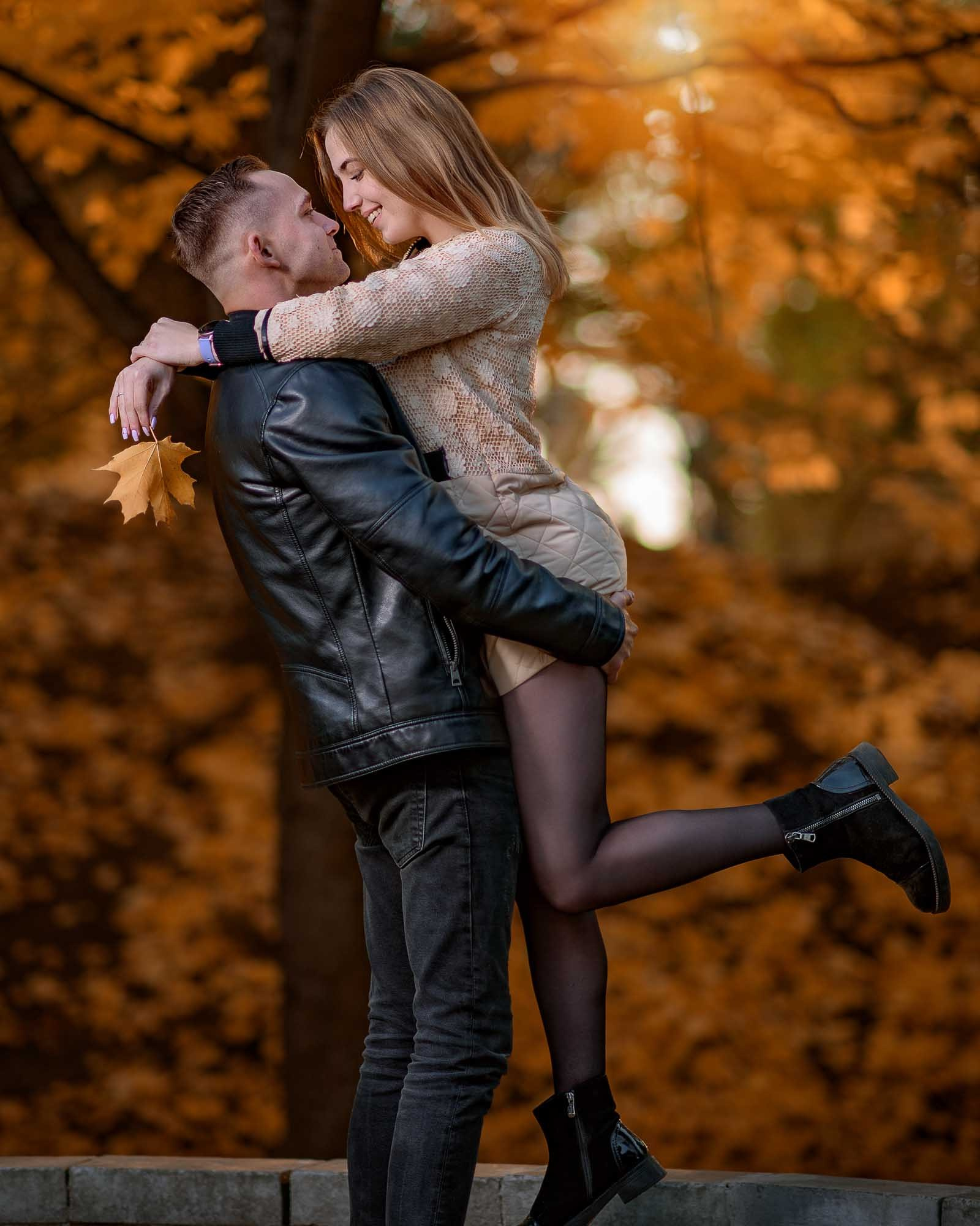 парень держит девушку на руках на фоне солнечного света и осенней листвы, в руках у девушки желтый лист клена