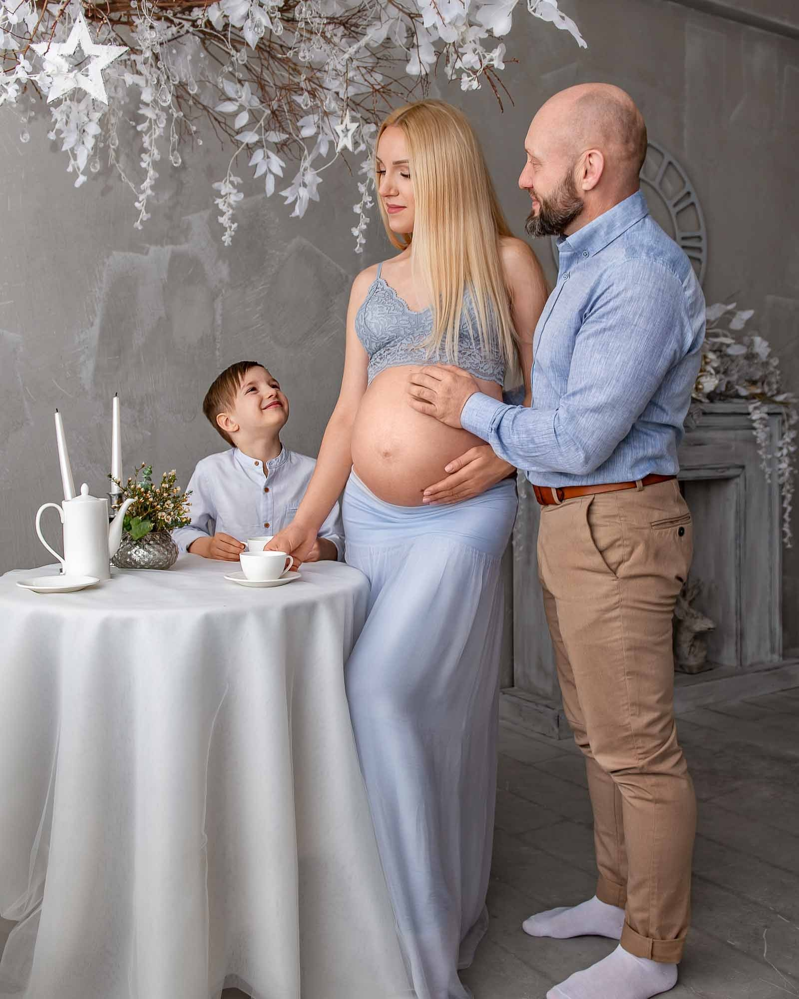 беременная женщина стоит около стола, ее обнимает муж, сын стоит рядом, на столе стоят свечи и кофе. Фото снято в студии города Борисова