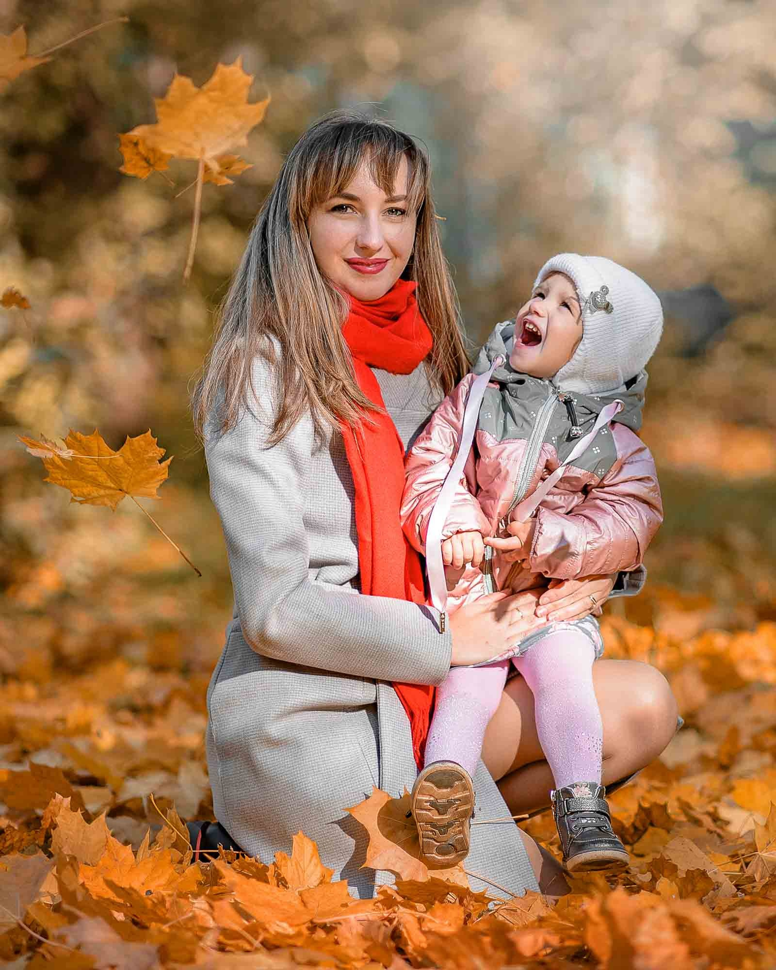 мама с дочкой улыбаются, вокруг яркая осенняя листва и солнечный свет