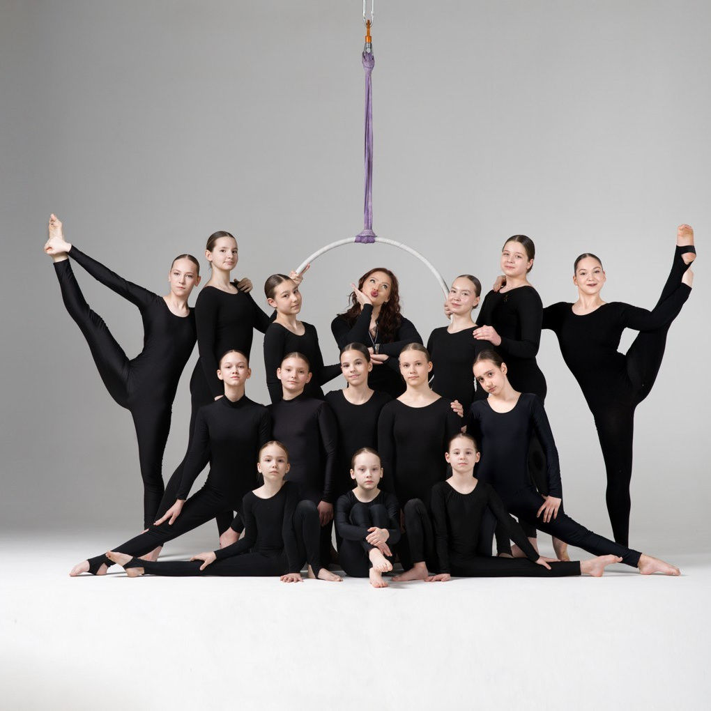 Групповое фото учениц и тренера школы воздушной гимнастики в Тюмени. Коллектив в черной тренировочной форме позирует в фотостудии на фоне воздушного кольца.