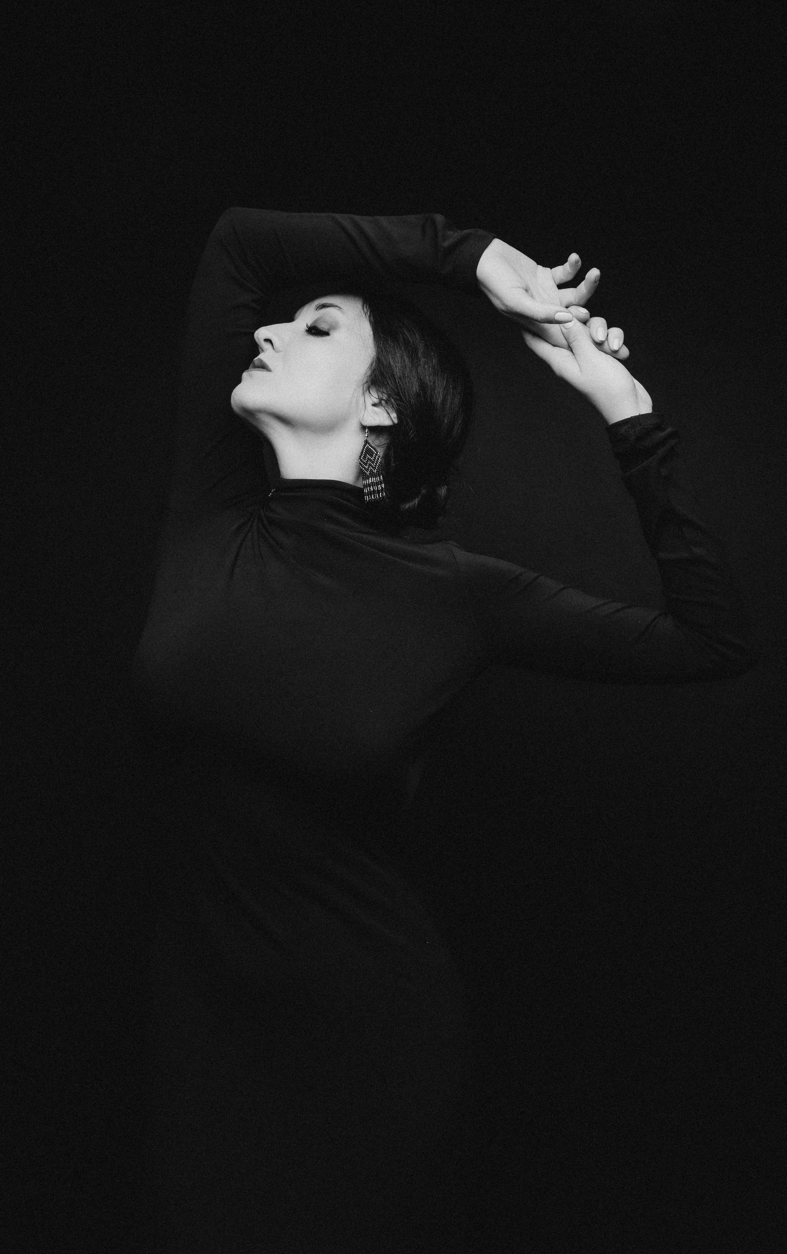 Женщина в черном обтягивающем платье с жемчужными серьгами, позирует с поднятыми руками на темном фоне.