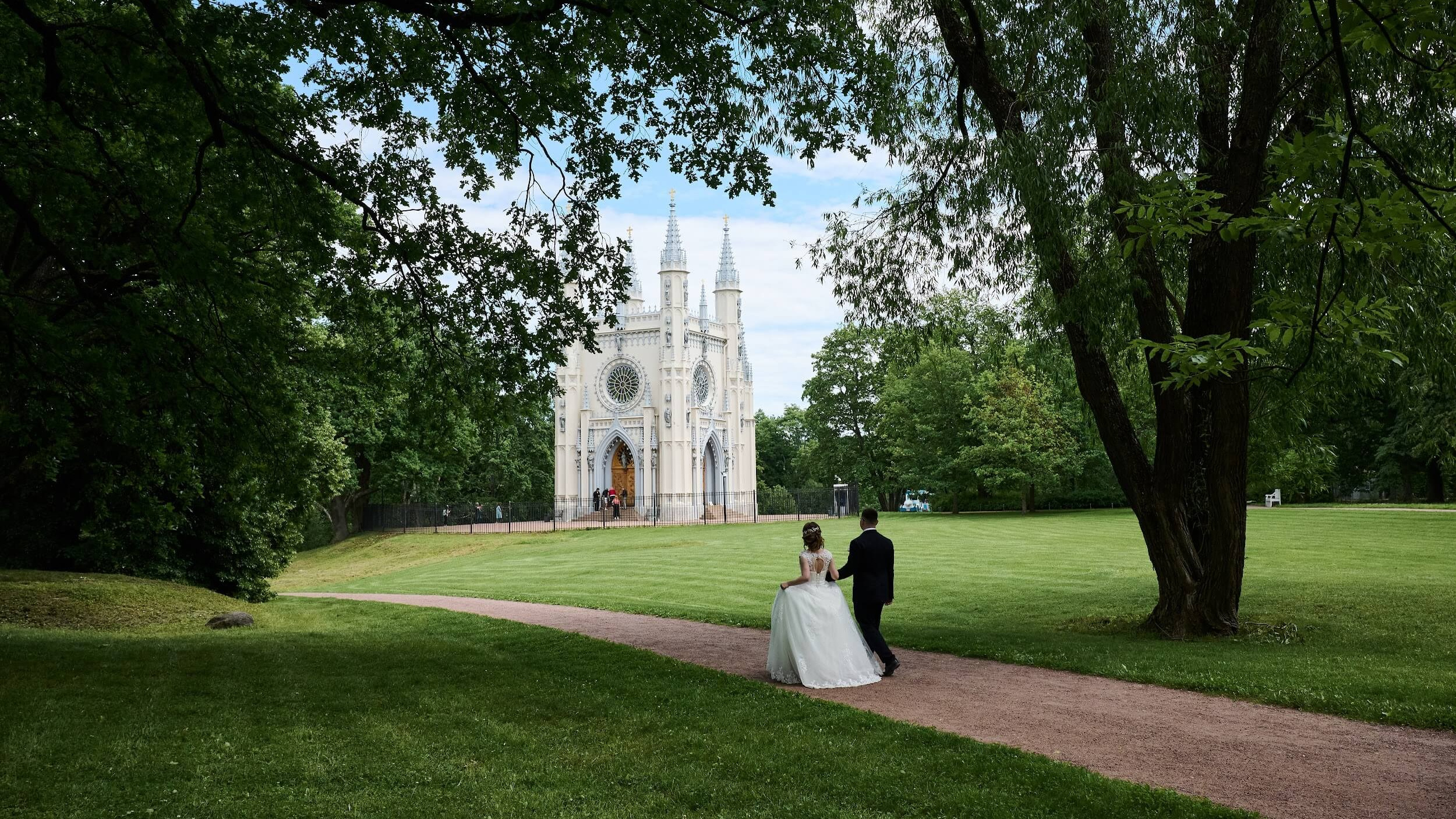 Молодожёны прогуливаются по аллеям парка Александрия. На заднем плане — изящная Готическая капелла. Естественный свет, зелёные насаждения. Пара держится за руки. Атмосфера спокойствия и романтики.