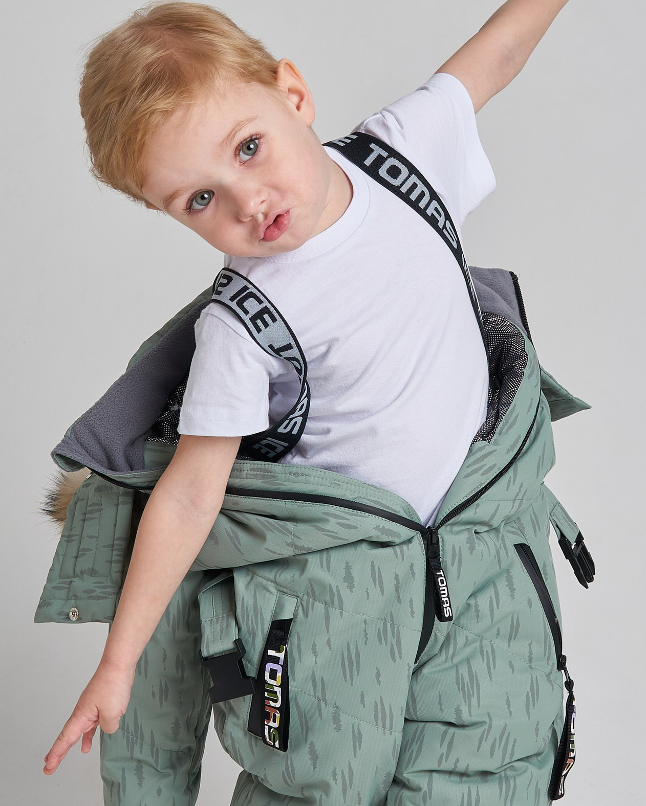 Мальчик в комбинезоне на сером фоне изображает руками самолет