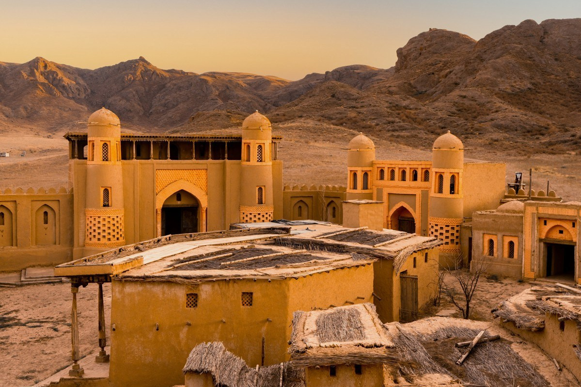 Пейзаж, напоминающий лунный, с извилистой рекой, окруженной густой зеленью и деревьями на фоне пустынных скалистых гор Лунного каньона. Солнечный день с ярко-голубым небом, украшенным небольшими облаками, подчеркивает природную красоту местности. Река Чарын образует небольшие островки, покрытые травой и кустарником.