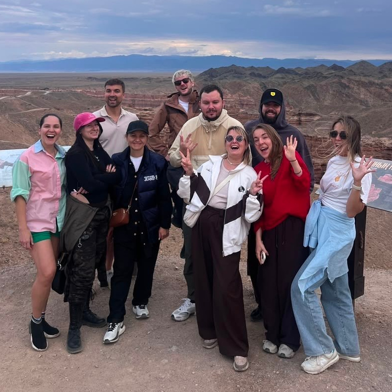 Группа людей, участников корпоративного тура Melon Travel на озеро Иссык, одетых в зимнюю одежду, позирует на фоне заснеженного пейзажа. На земле лежит снег, деревья покрыты инеем, а на заднем плане виднеется туман, создающий атмосферу спокойствия и загадочности. Участники группы улыбаются и выглядят счастливыми.