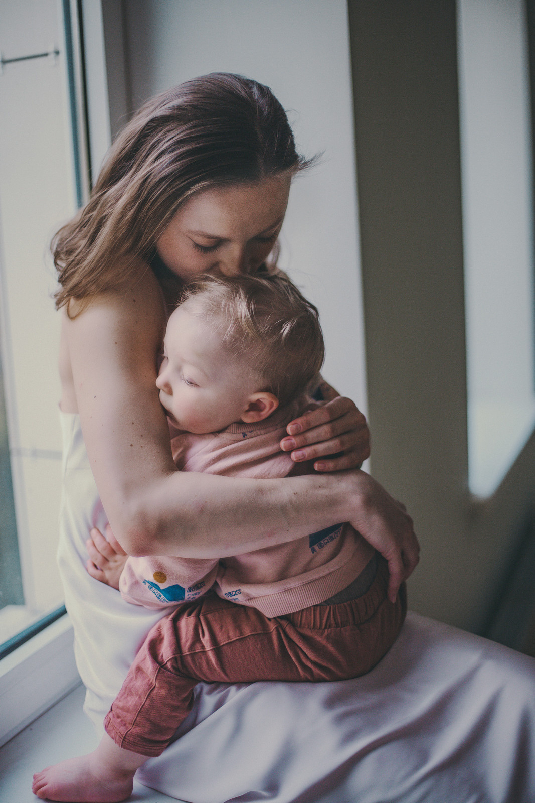 молодая мама с малышом на руках сидят на окне в обнимку, мама целует сына в макушку
