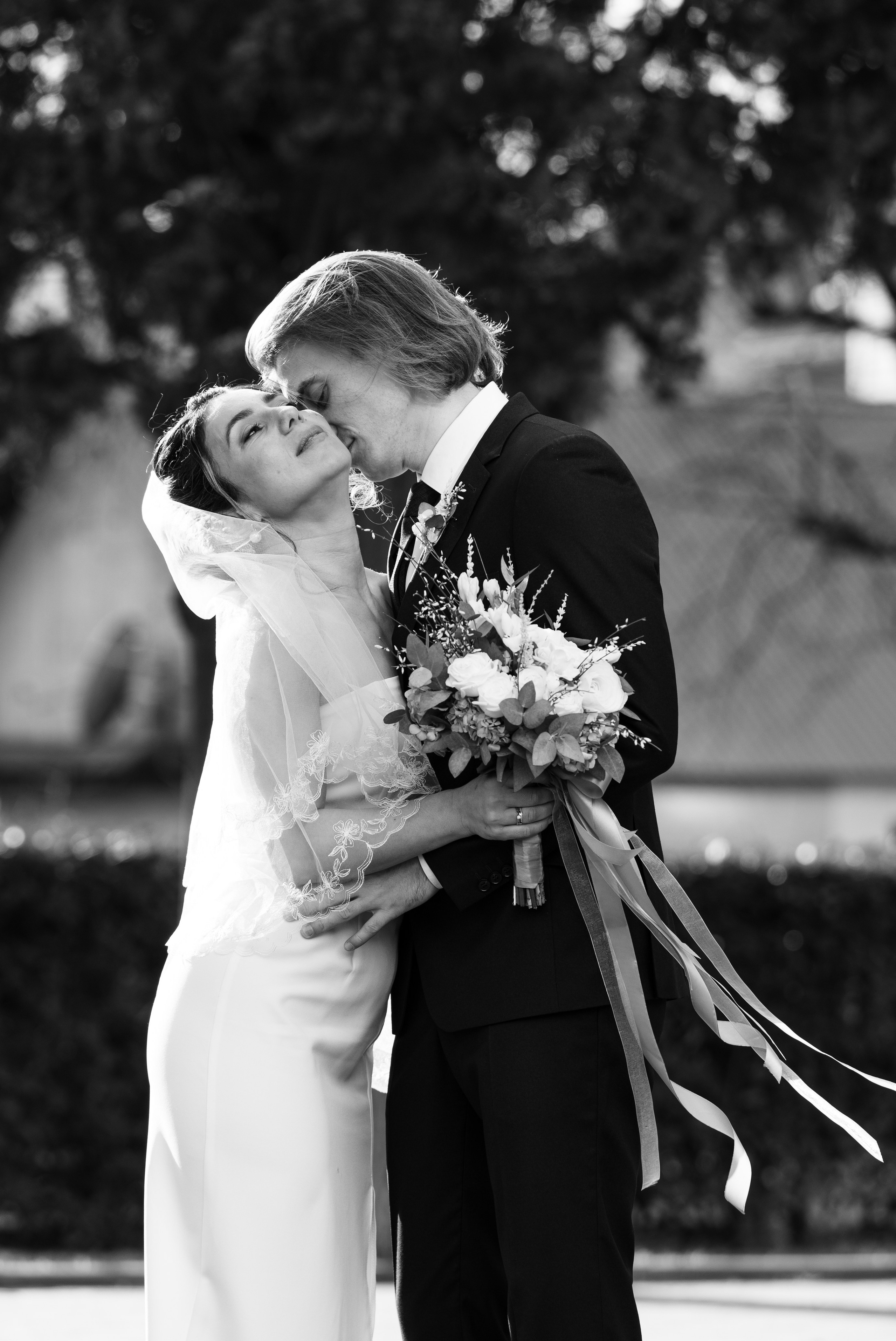 Это черно-белая фотография жениха и невесты в день их свадьбы. Невеста одета в белое платье с длинной вуалью. Жених одет в черный костюм с белой рубашкой и черным галстуком. Невеста держит букет цветов. Жених целует невесту в лоб. Они стоят в парке на фоне деревьев и кустарников.