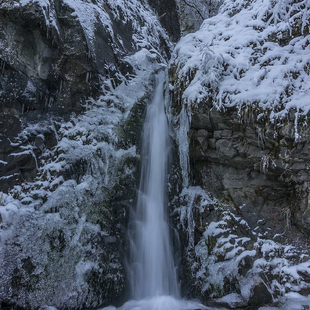 Человек стоит у подножия замерзшего водопада Медвежий среди заснеженных скал. Вода продолжает течь, частично покрытая льдом, создавая красивый контраст между движущейся водой и ледяными наростами. Земля вокруг покрыта снегом, создавая зимнюю атмосферу. Человек одет в теплую одежду — синее пальто с капюшоном и розовую шапку.