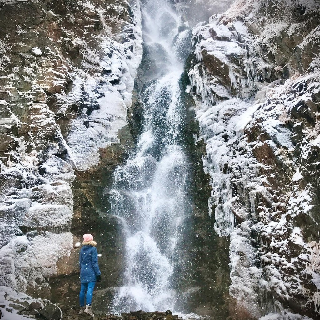 Человек стоит у подножия замерзшего водопада Медвежий среди заснеженных скал. Вода продолжает течь, частично покрытая льдом, создавая красивый контраст между движущейся водой и ледяными наростами. Земля вокруг покрыта снегом, создавая зимнюю атмосферу. Человек одет в теплую одежду — синее пальто с капюшоном и розовую шапку.