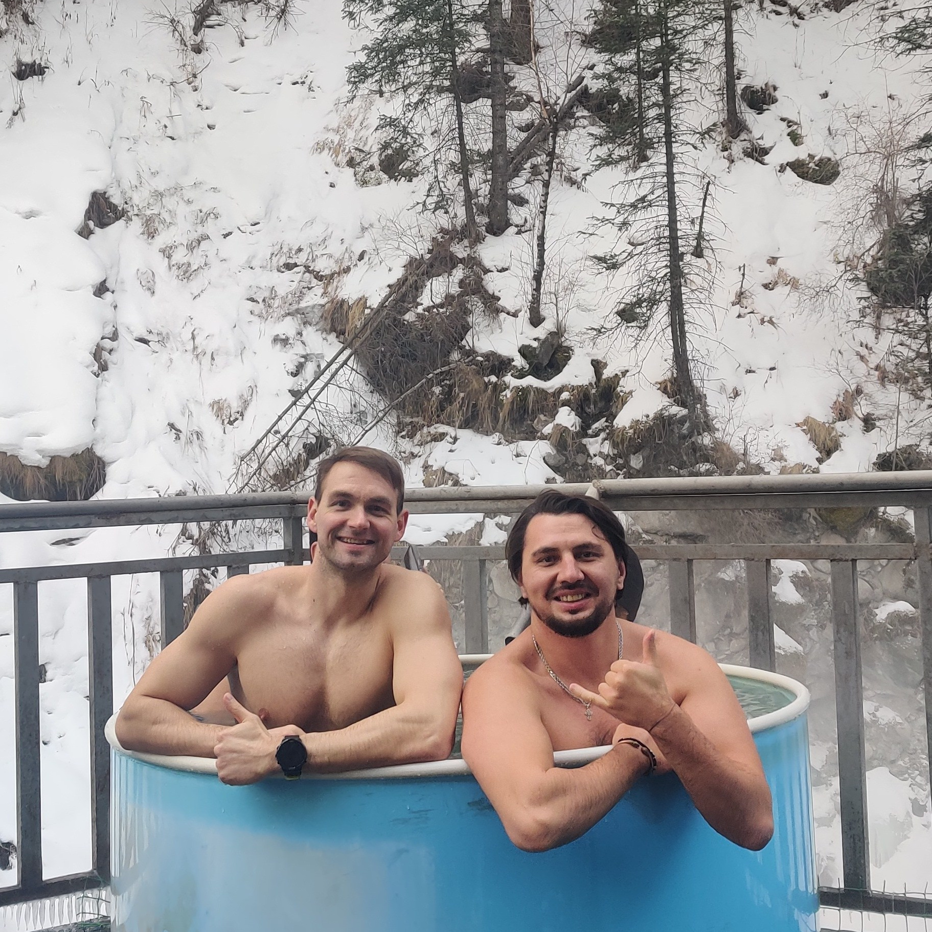 Человек стоит у подножия замерзшего водопада Медвежий среди заснеженных скал. Вода продолжает течь, частично покрытая льдом, создавая красивый контраст между движущейся водой и ледяными наростами. Земля вокруг покрыта снегом, создавая зимнюю атмосферу. Человек одет в теплую одежду — синее пальто с капюшоном и розовую шапку.