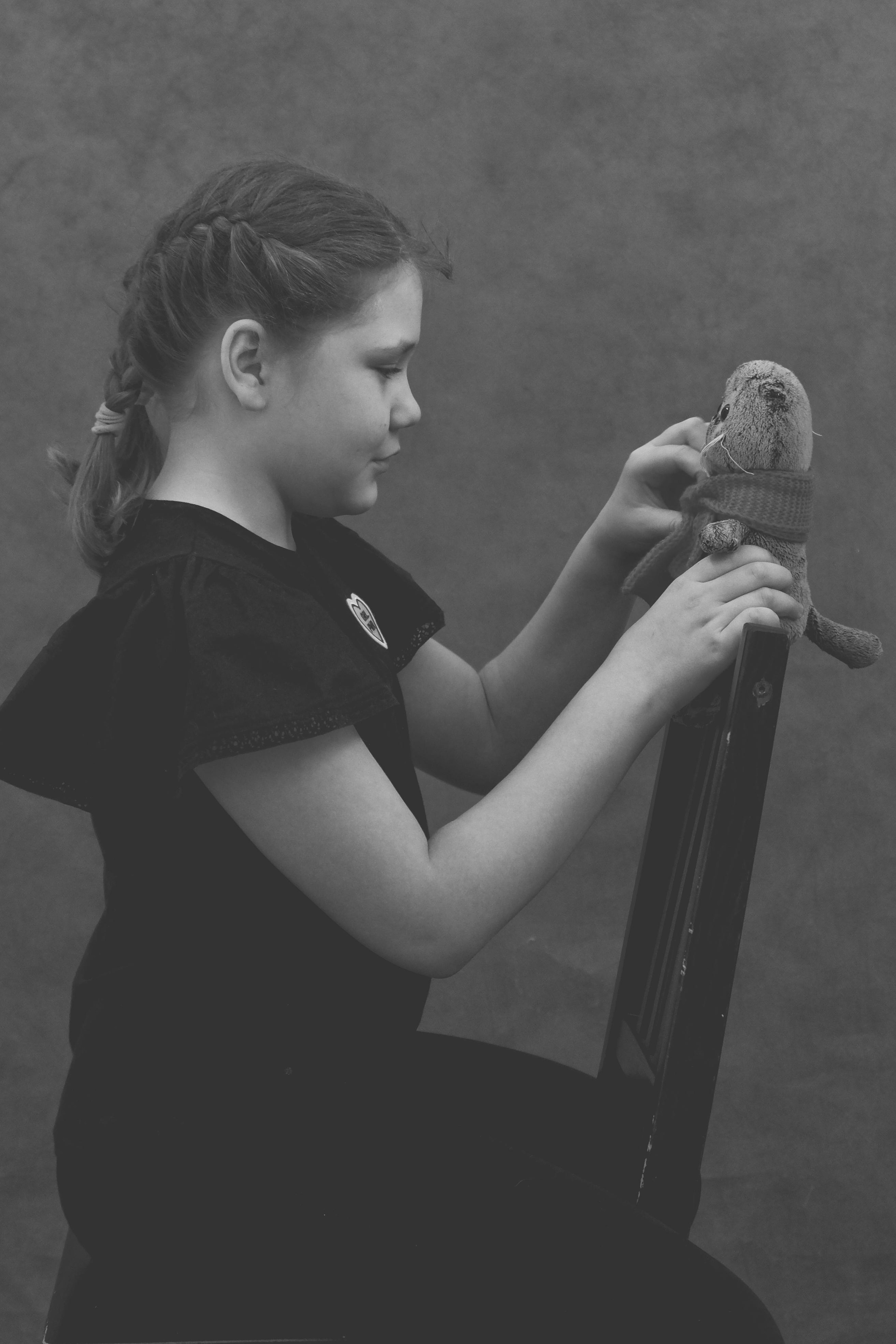 девочка на стуле положила руки на спинку стула и держит игрушечного кота басика 