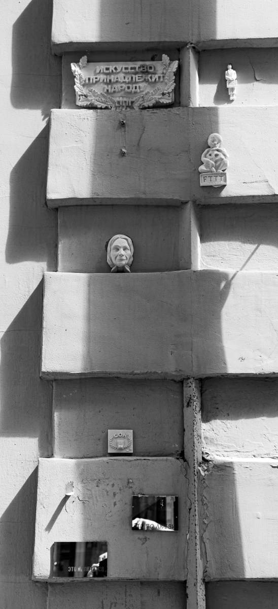 Черно-белая архитектурная фотография: стена здания с вертикальным рустом, в нишах размещены табличка "Искусство принадлежит народу", гипсовые фигурки (бюст женщины, ребенок с игрушкой, маленькая человеческая фигурка), внизу зеркальные наклейки с надписями "Это не Ленин", "А по деревьям", городская память, археология душ, уличная фотография Москвы, Вадим Топорёнок, Манифест теней, конфликт эпох.
