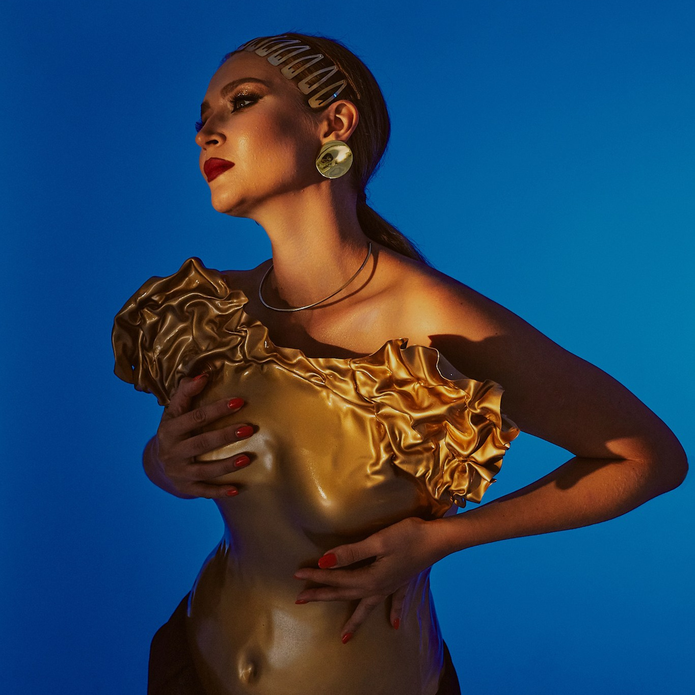 Авангардная фотография: модель в необычном золотом топе и черных брюках позирует на синем фоне, подчёркивая смелый и эксцентричный образ.