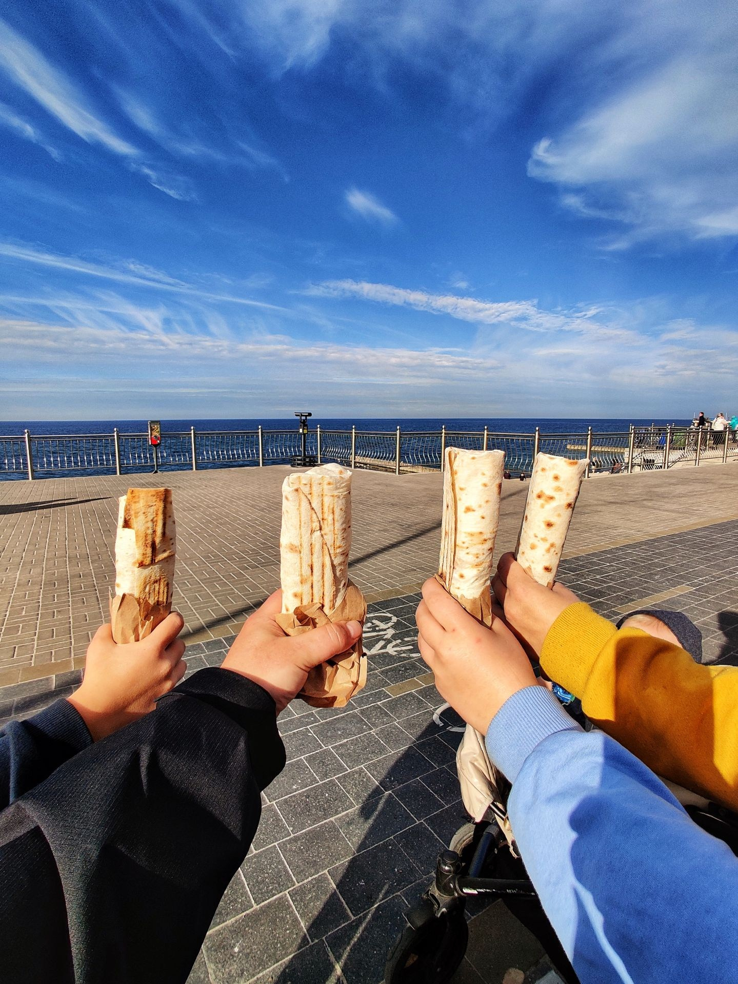 Четыре руки держат шаурму на набережной с видом на море и ясное голубое небо над горизонтом