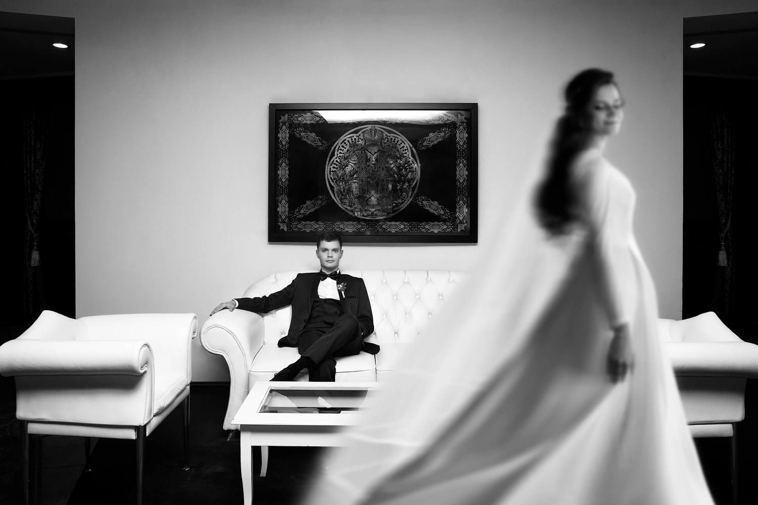 Жених сидит на диване в лаунж-зоне отеля и невеста проходит перед ним, чб