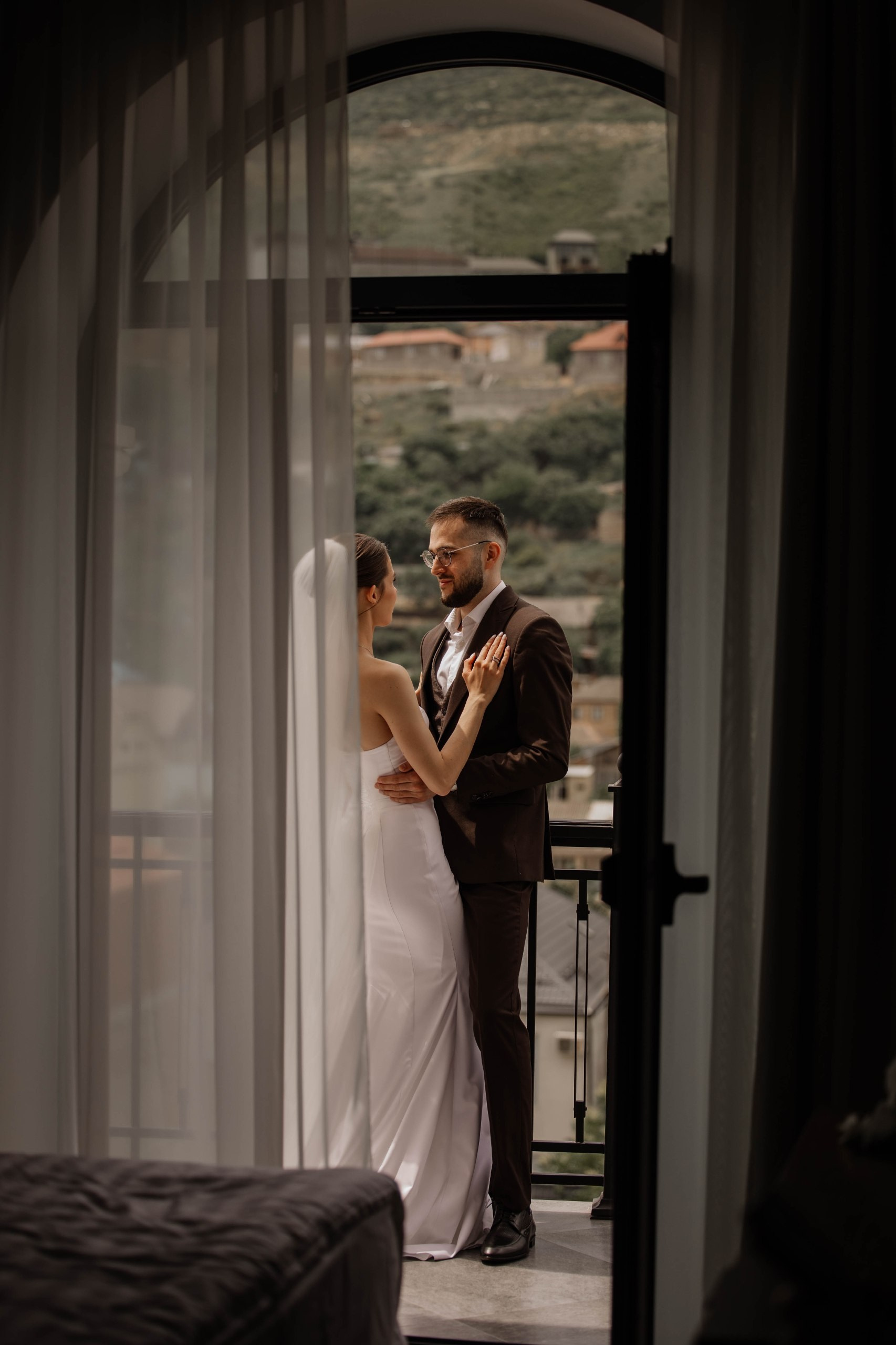 Свадебные фотографии у окна рядом с горами, жених обнимает невесту