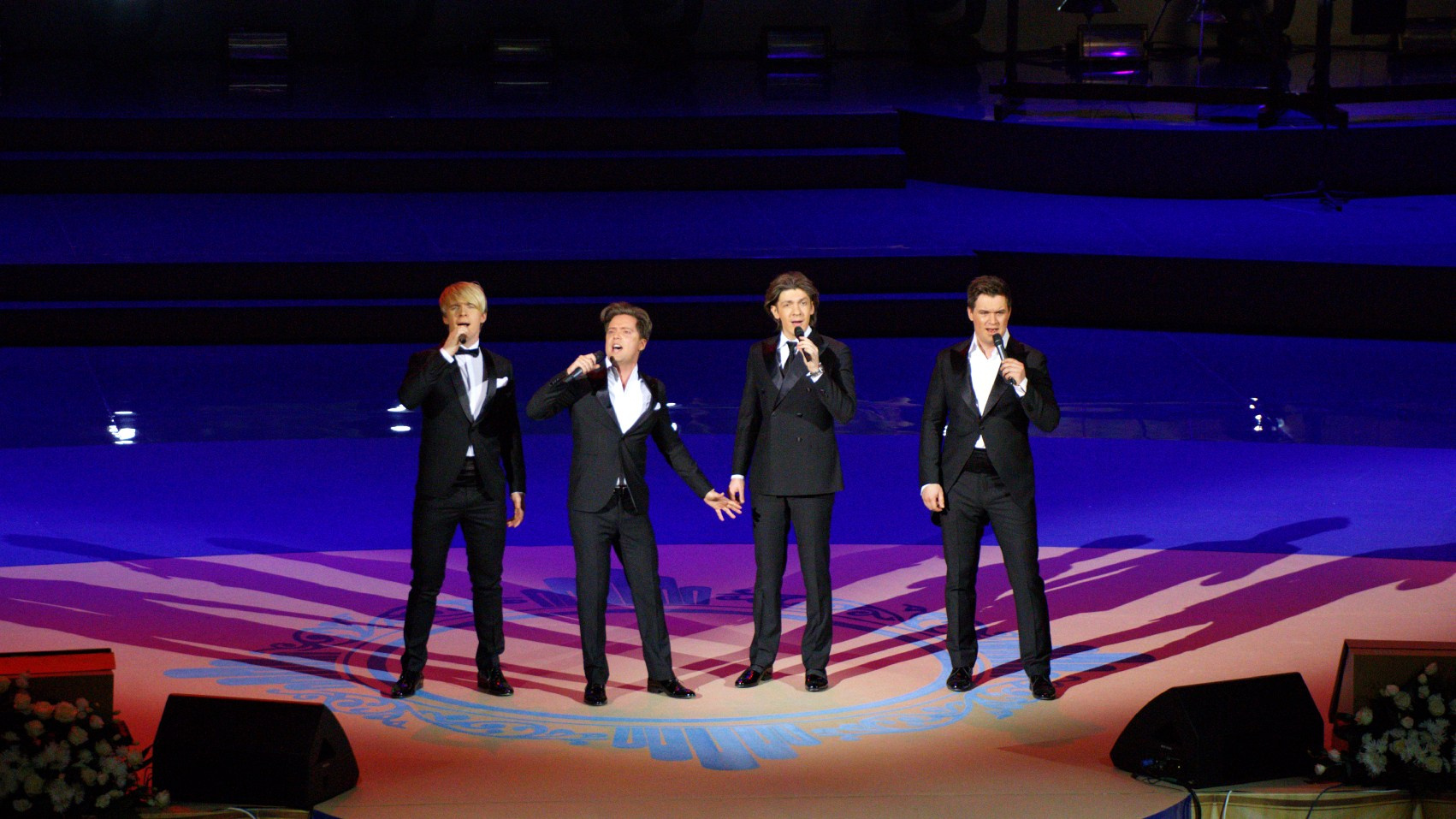 Вокальный квартет - четыре исполнителя в черных костюмах выступают на сцене с микрофонами, концертная фотография