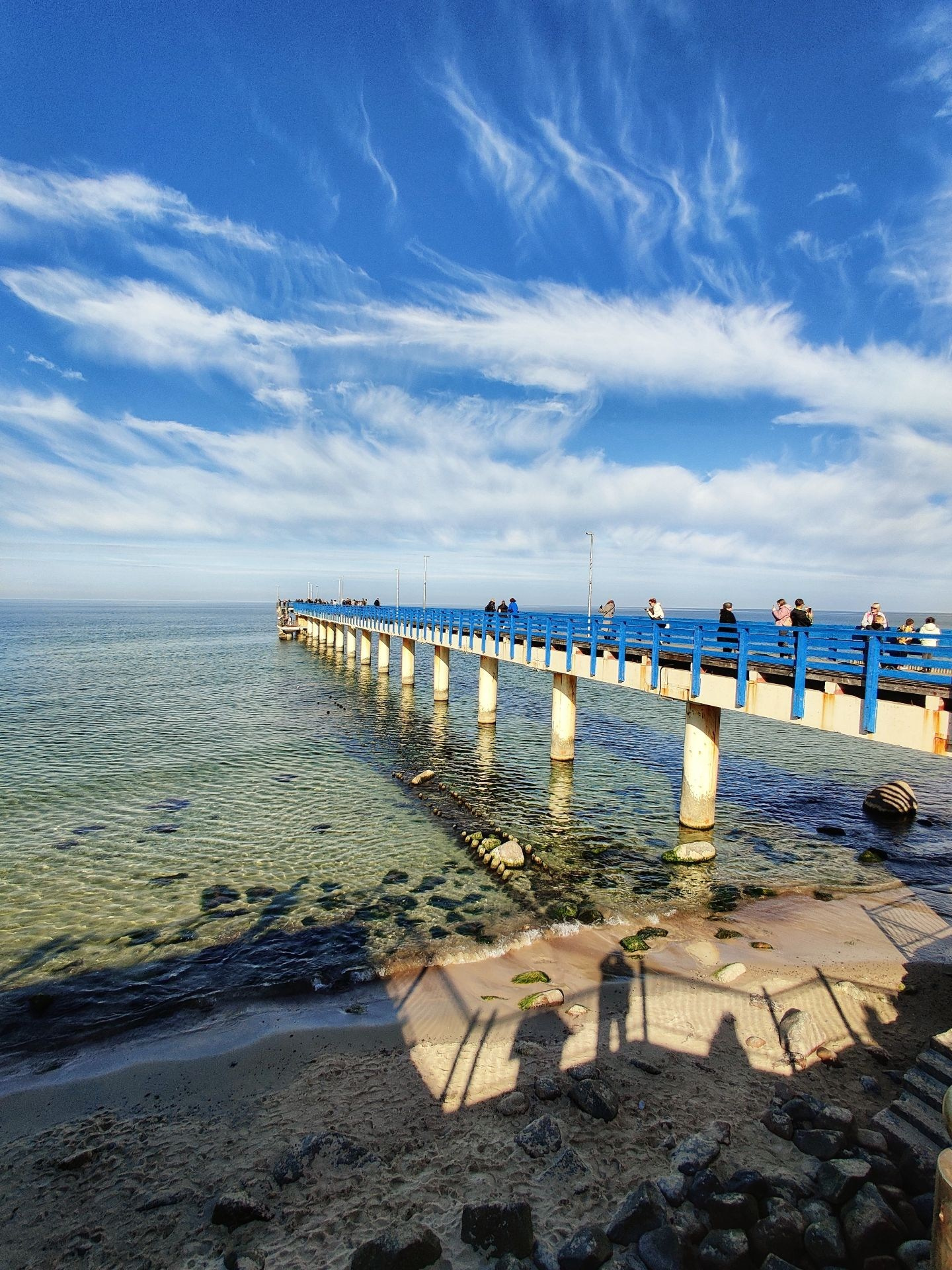 Длинный пирс с синими перилами, уходящий в спокойное море под живописными облаками на пляже Зеленоградска
