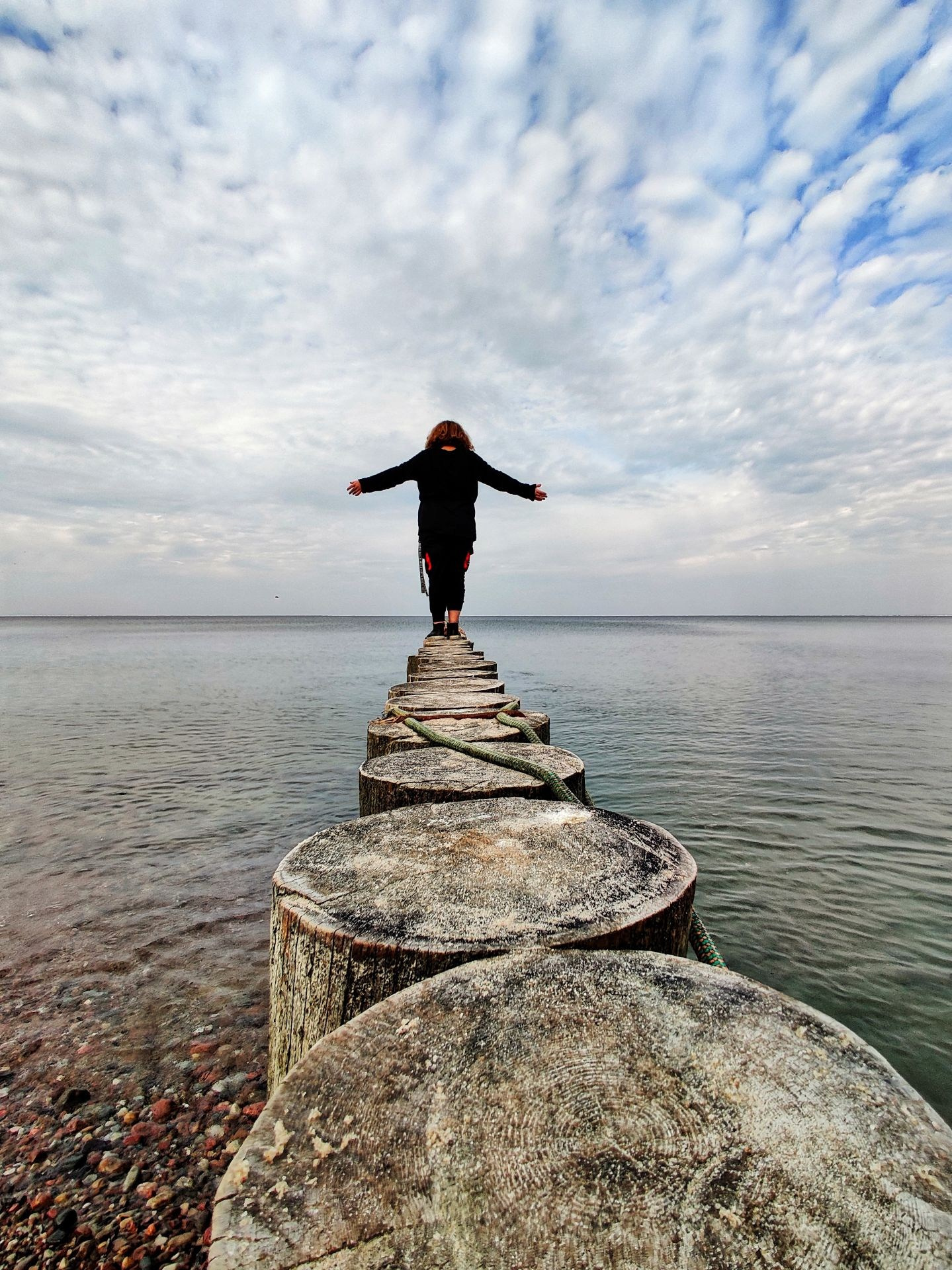 Человек с раскинутыми руками идёт под облачным небом по деревянным сваям, уходящим в море