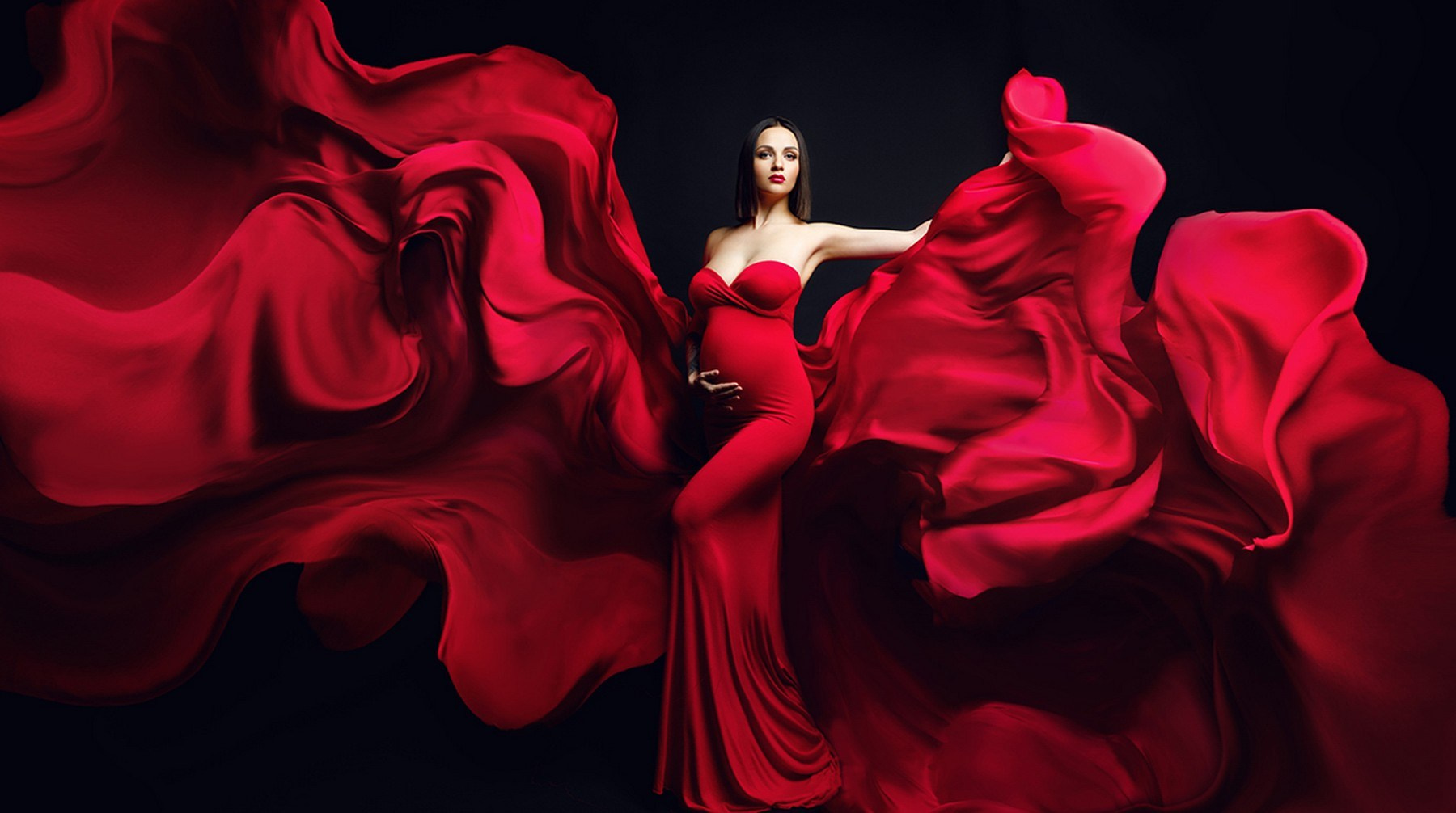 Беременная девушка в красном платье с летящей тканью позирует на фоне.