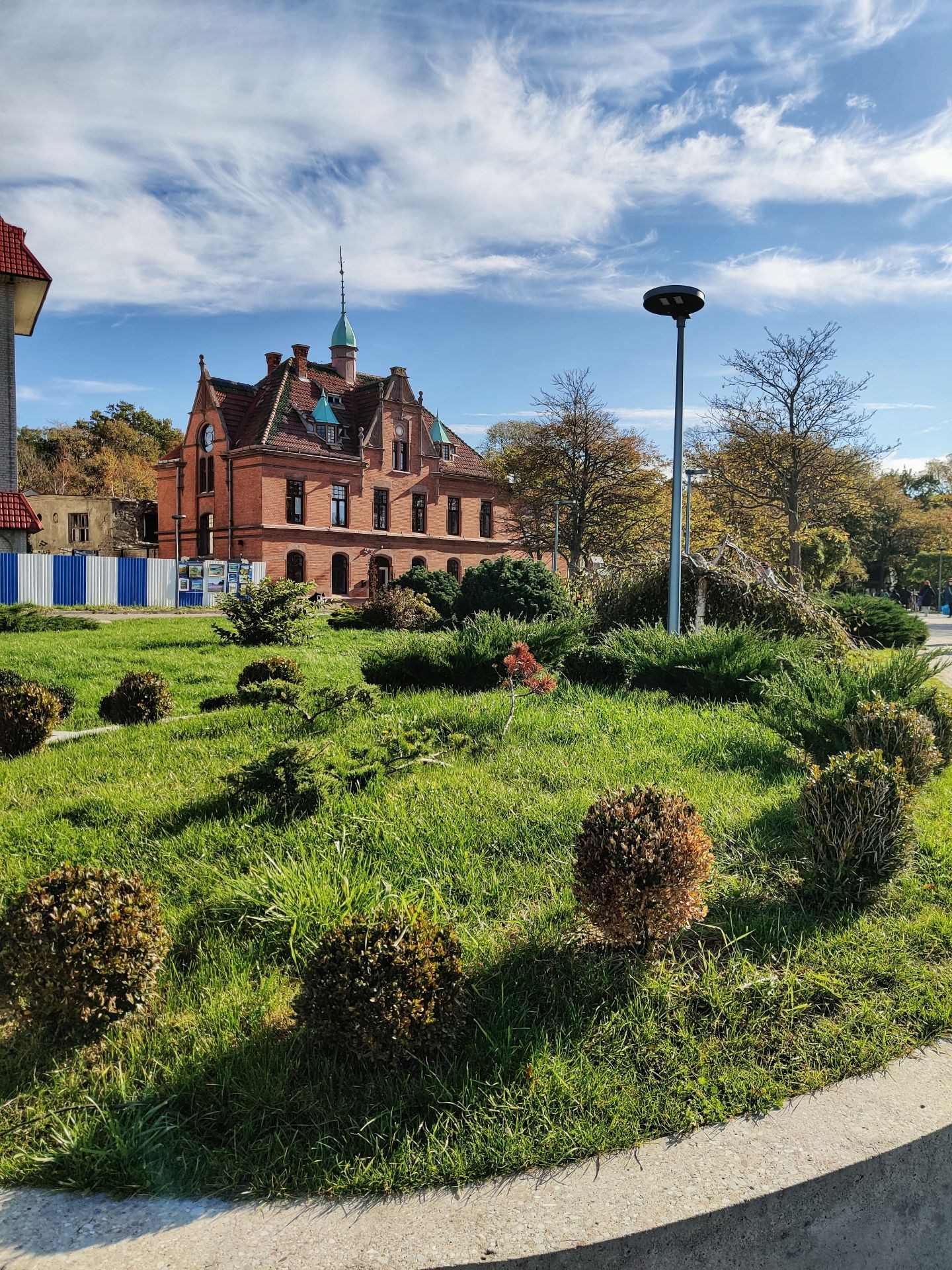 Историческое кирпичное здание с башенками на фоне голубого неба и ухоженного круглого газона с декоративными кустами
