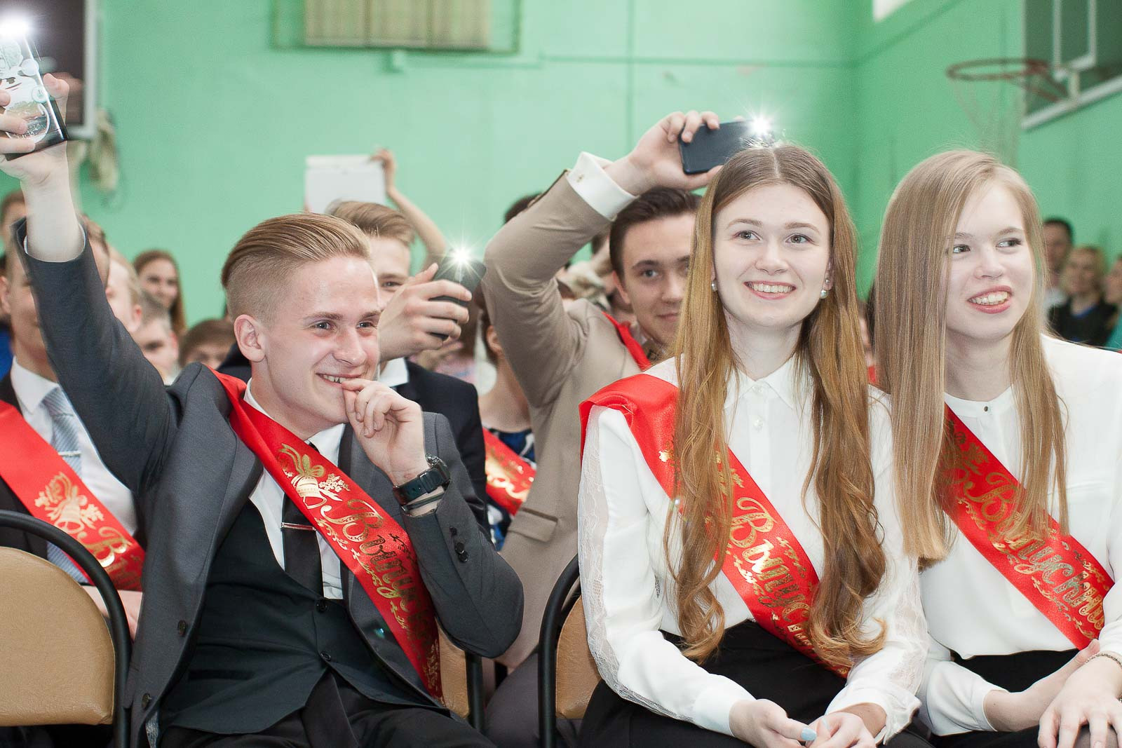 Выпускники 11 класса с лентами выпускника сидят на стульях и улыбаются.