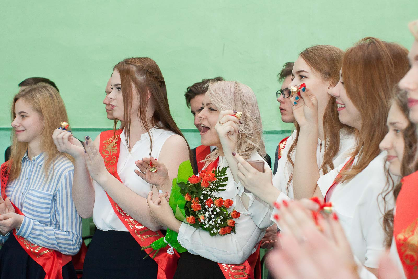 Выпускницы 11 класса аплодируют стоя на фоне зеленой стены.