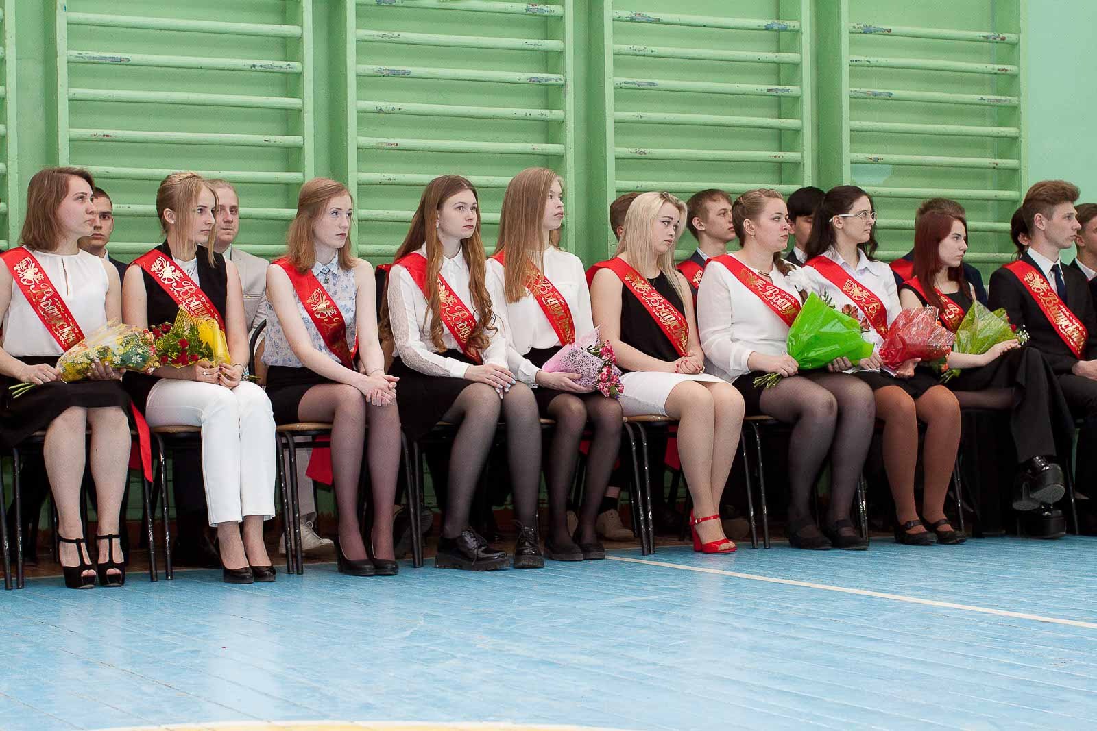 Выпускники 11 класса с красными лентами сидят вдоль шведской стенки в школьном спортзале.