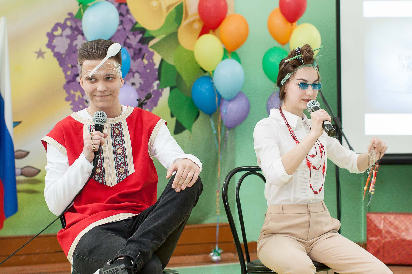 Юноша и девушка играют сценку сидя на стульях на фоне разноцветных шаров.