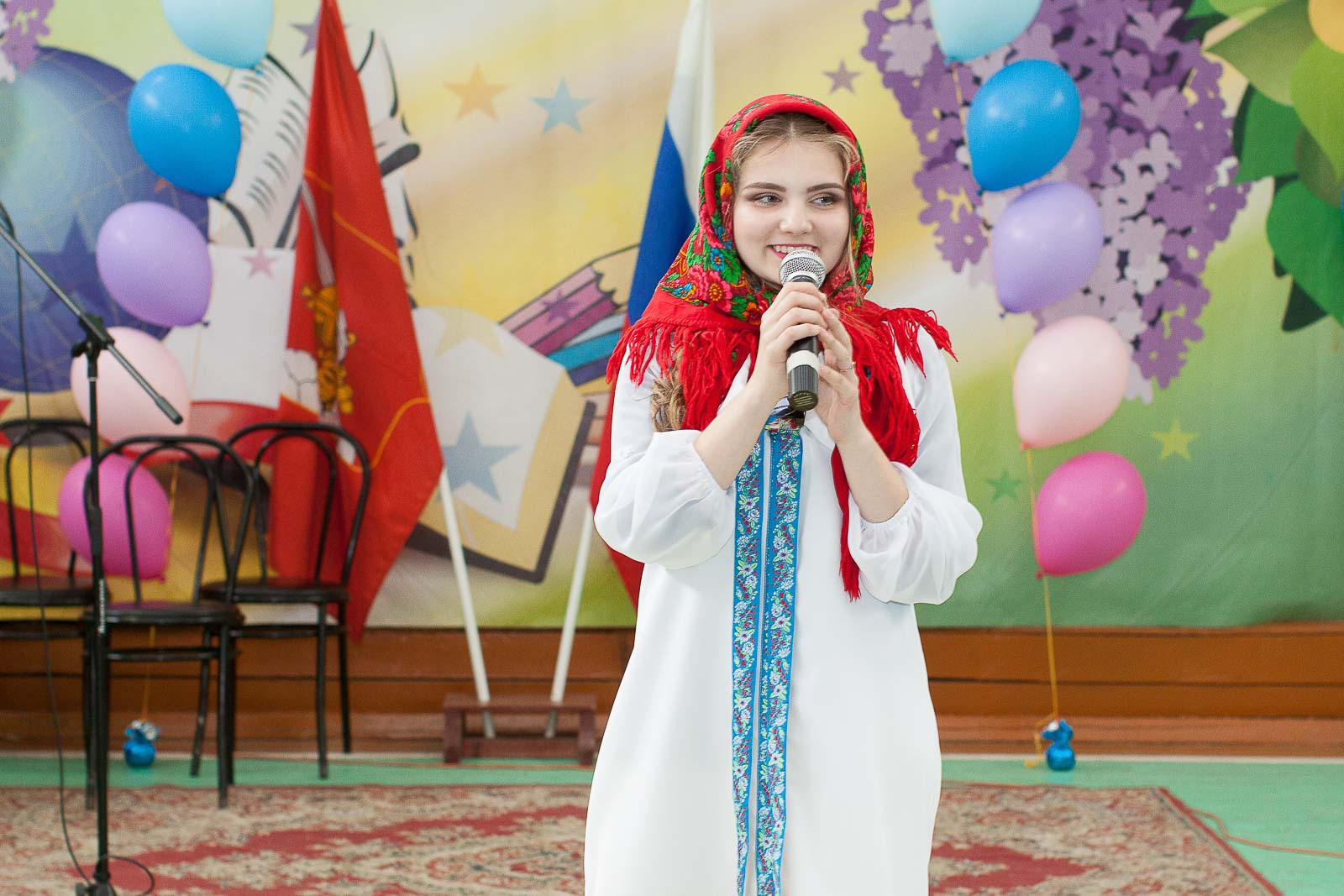 Девушка в красном платке и белом платье говорит в микрофон в школьном спортзале.