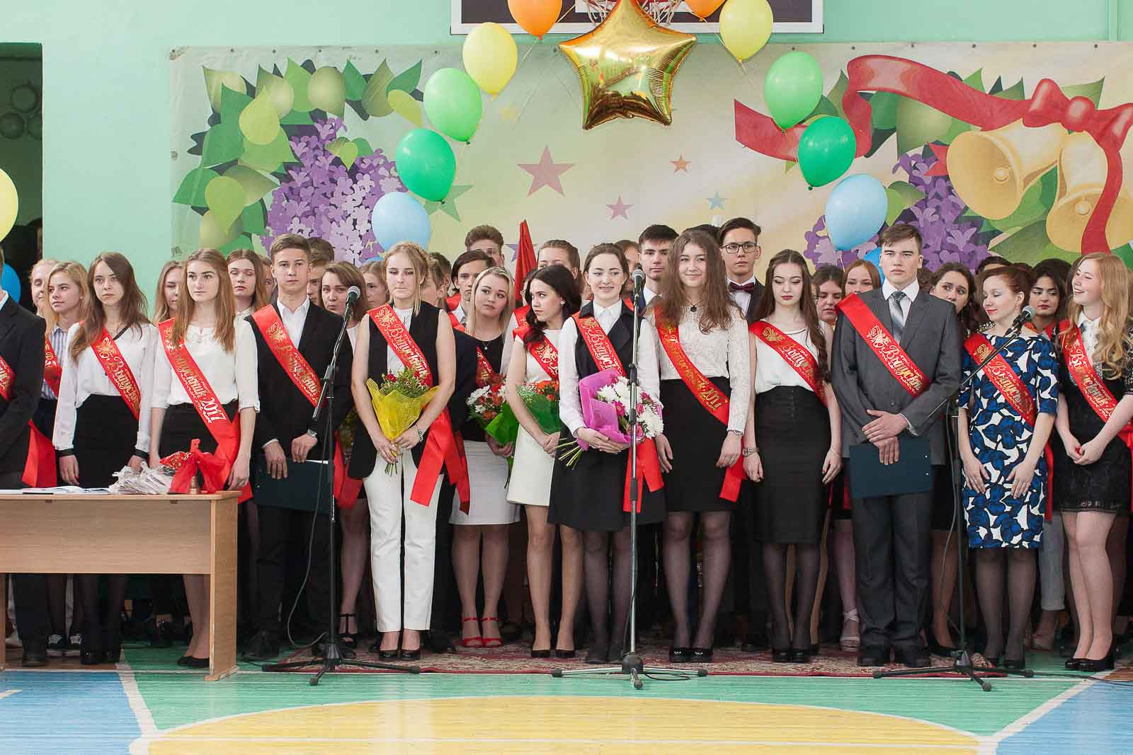 Выпускники 11 класса поют стоя хором песню на последнем звонке в спортзале.