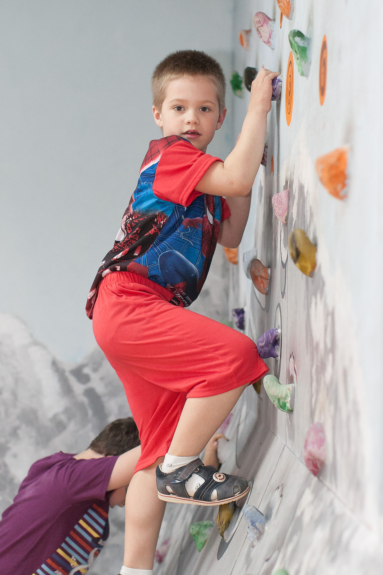 Мальчик в красных брюках забирается по стене скалодрома.