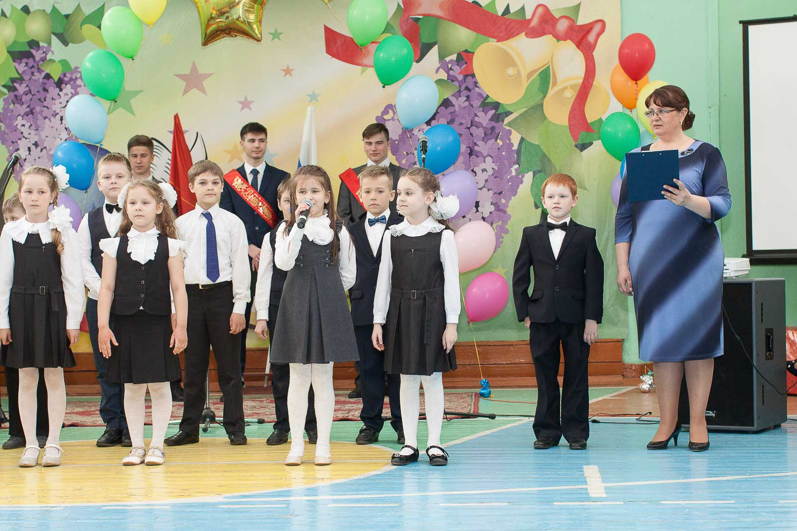 Первоклассники в парадной форме читают стихотворение в украшенном школьном спортзале.